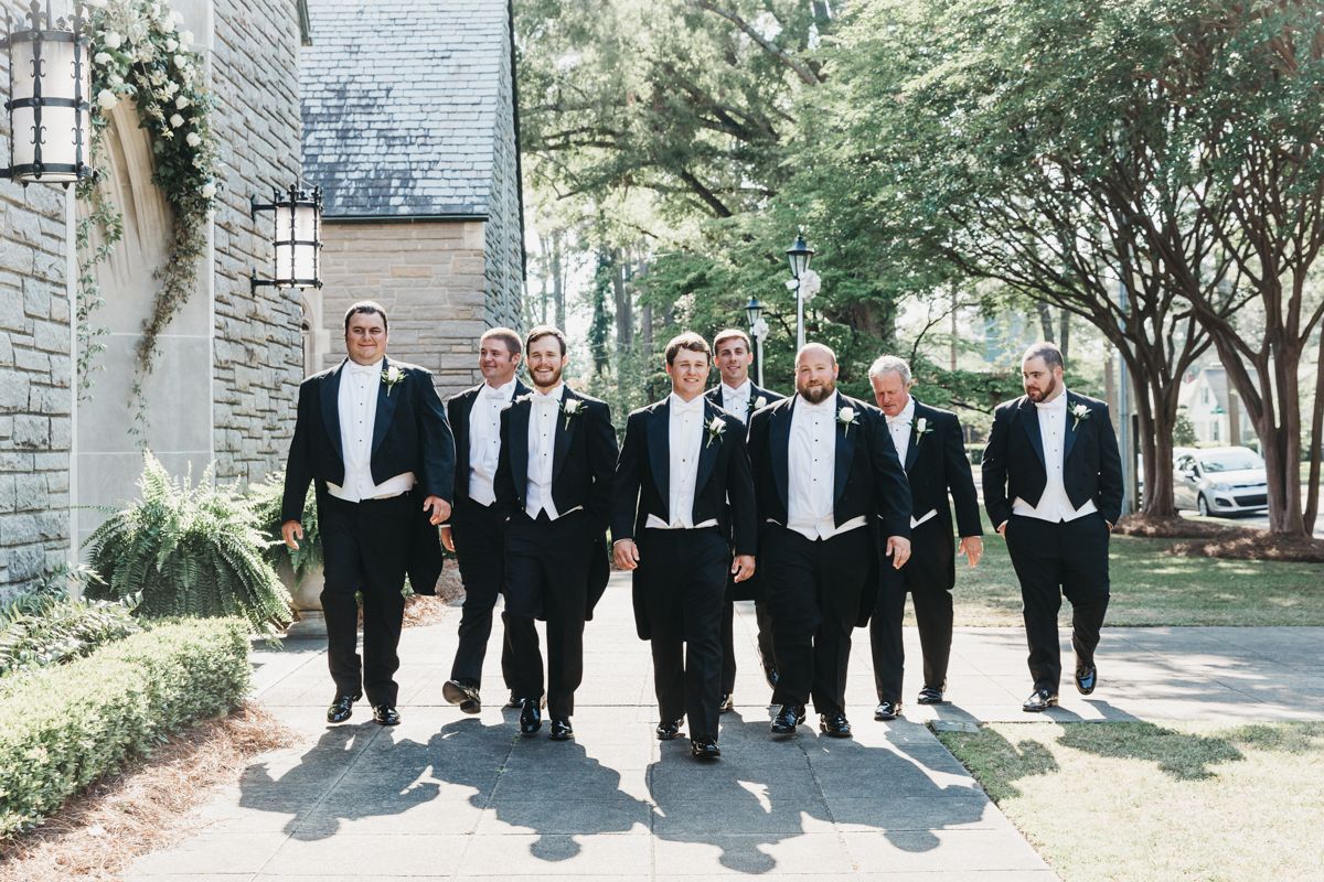 Groomsmen in formal attire walking along stone path surrounded by greenery.