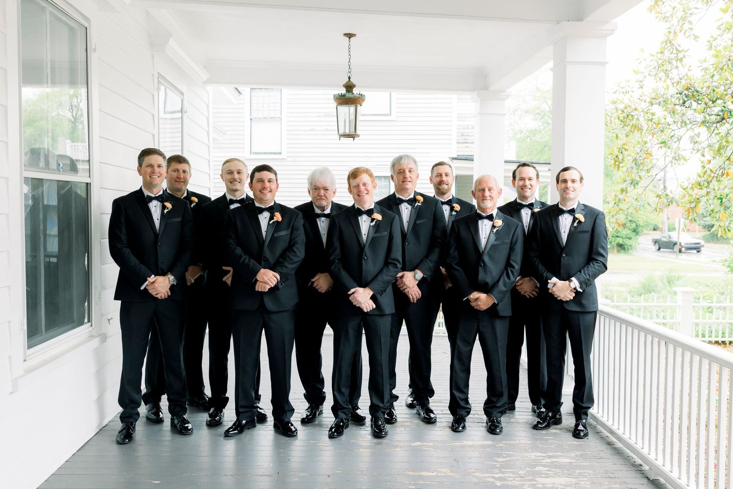 Groomsmen in black tuxedos with bow ties posing on a white porch for a wedding photo.