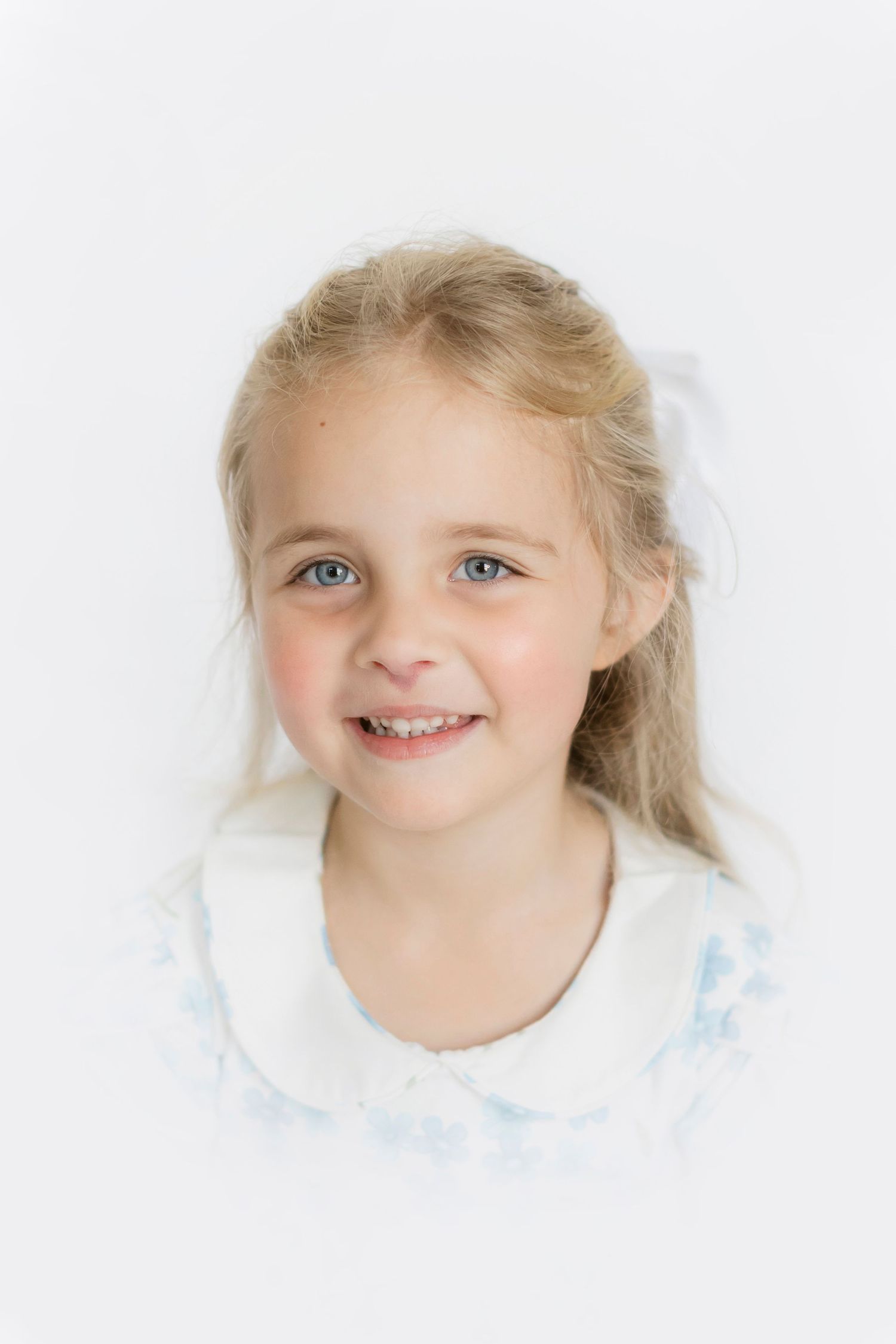 Child wearing white collared dress smiling brightly against light background.