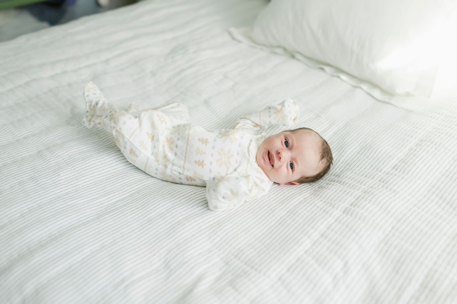 A smiling baby in white patterned clothing lies on a textured white bedspread.