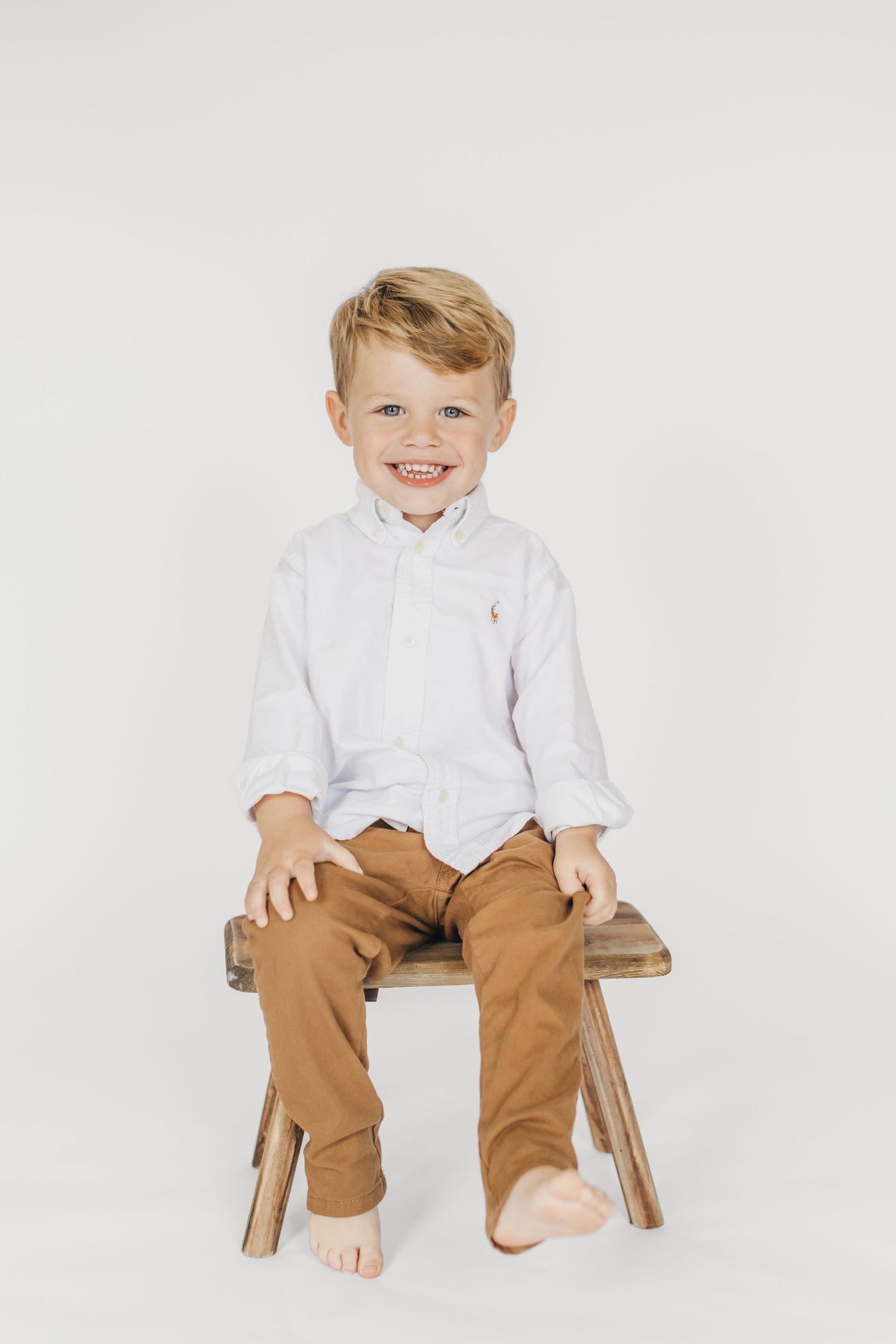 Young child sitting on wooden stool wearing white shirt and brown pants against white background.