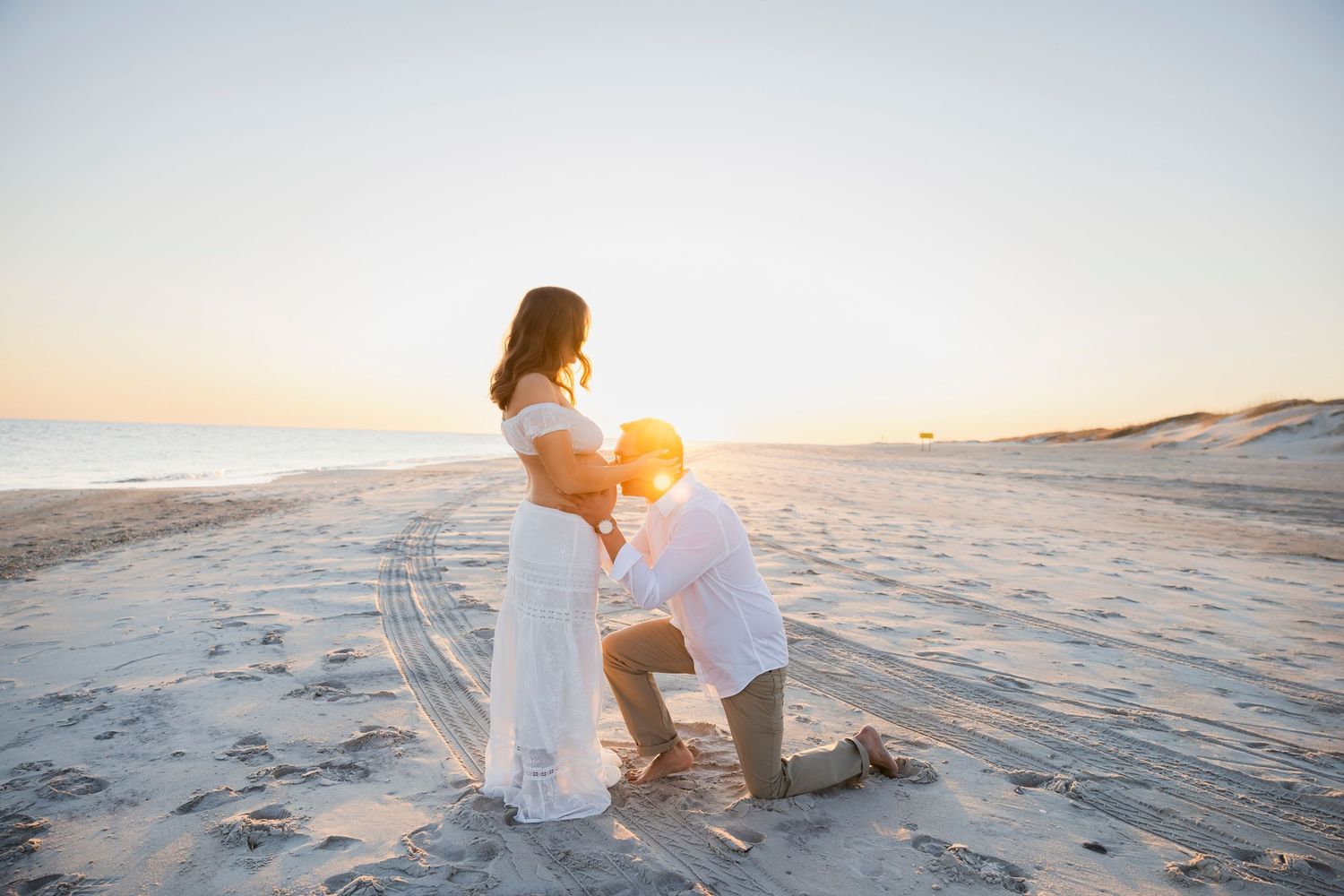 A couple sits together on a beach at sunset with ocean waves in the background.