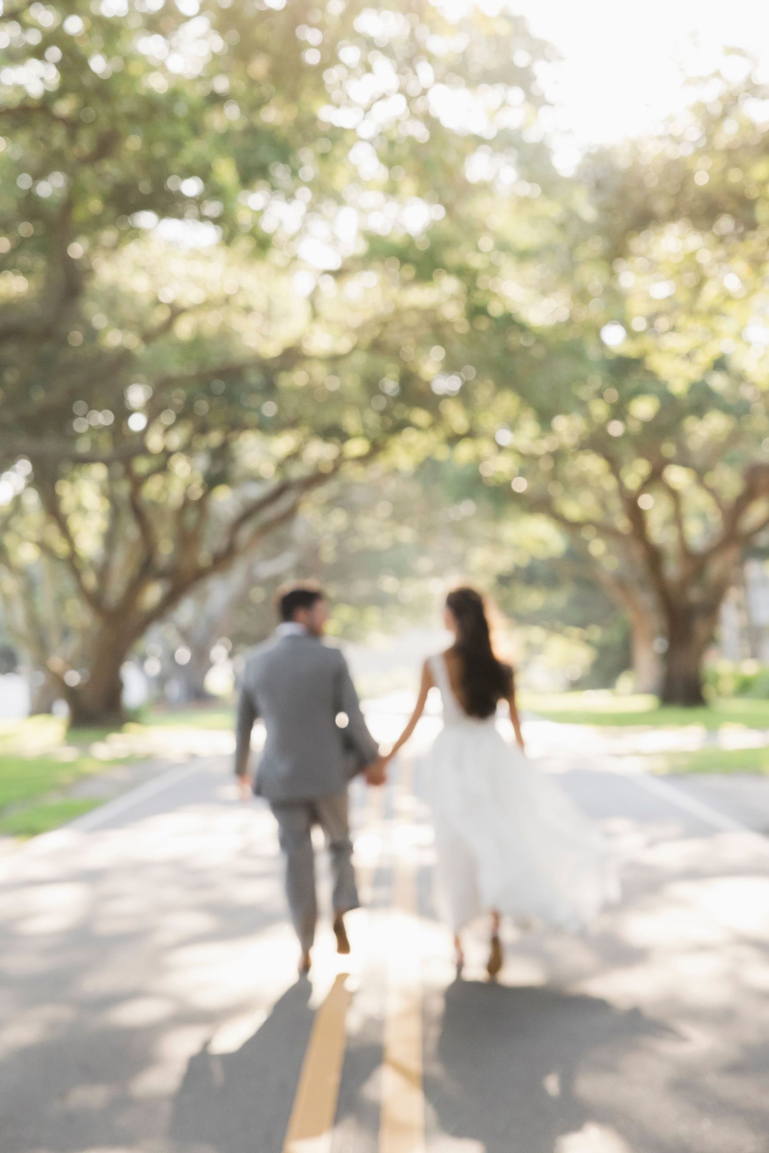 Blurred photo of a bride and groom holding hands walking down a tree-lined road on their wedding day.