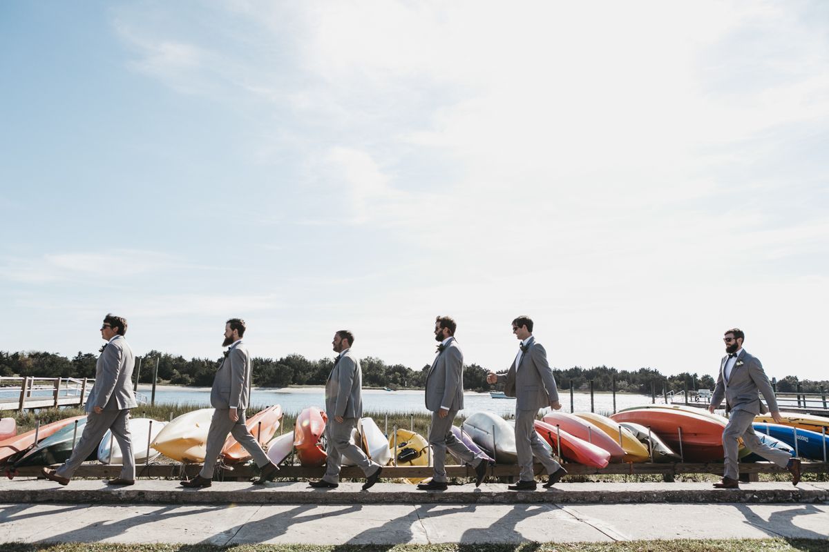 Wedding party members walk in line across a wooden boardwalk with water views.