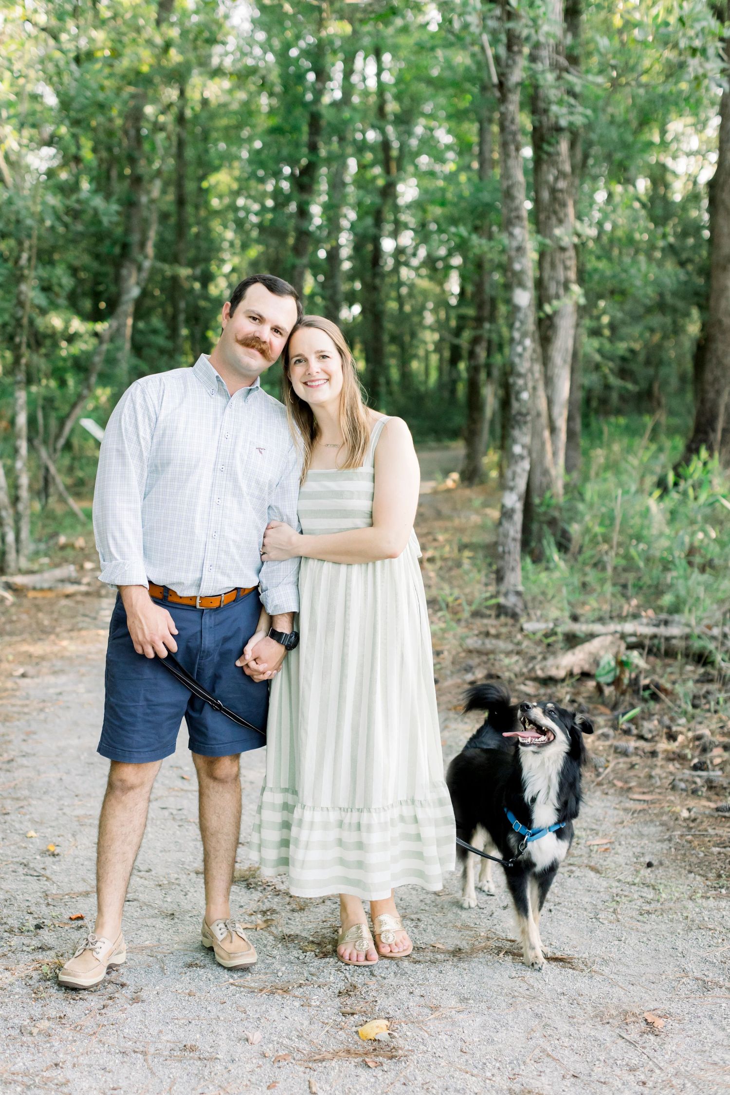 A casual couple poses on a wooded path with their border collie dog.