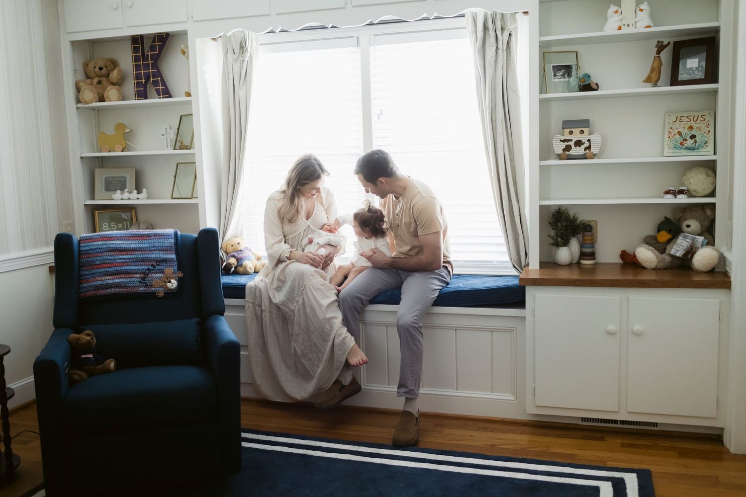 A family sits together on a window seat in a nursery filled with shelves of toys.