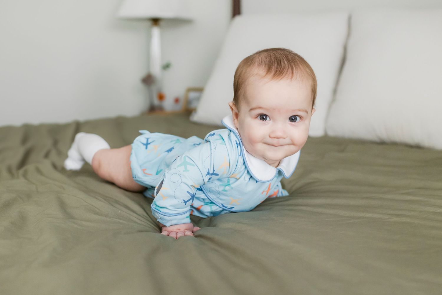 Baby in light blue outfit smiles while lying on gray bedding during indoor photo session.