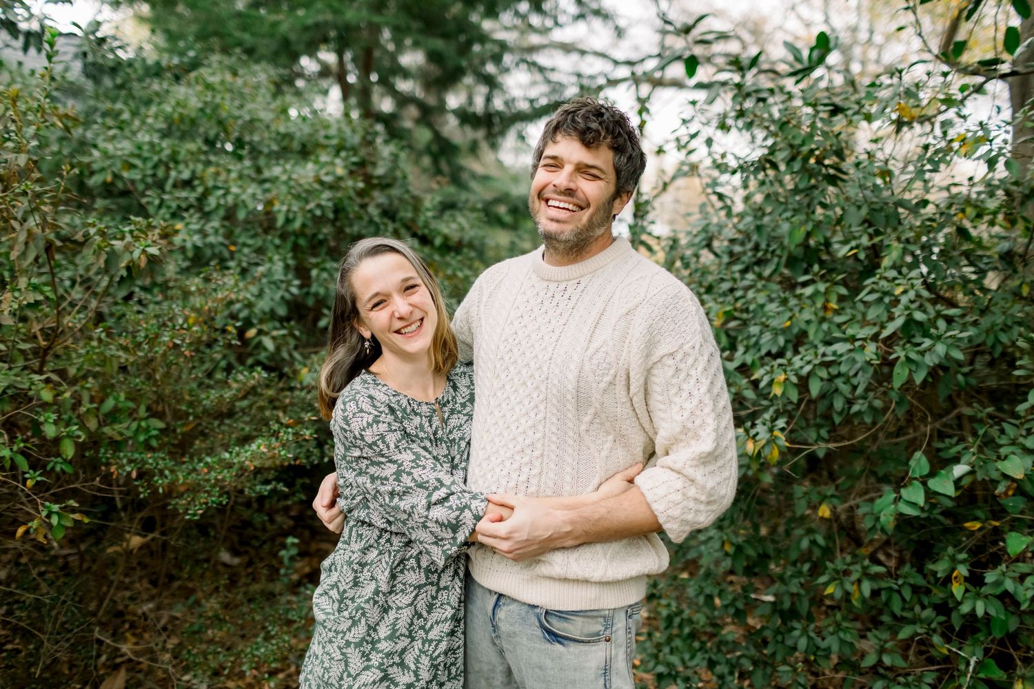 Couple laughing together outdoors in casual winter sweaters among green foliage.