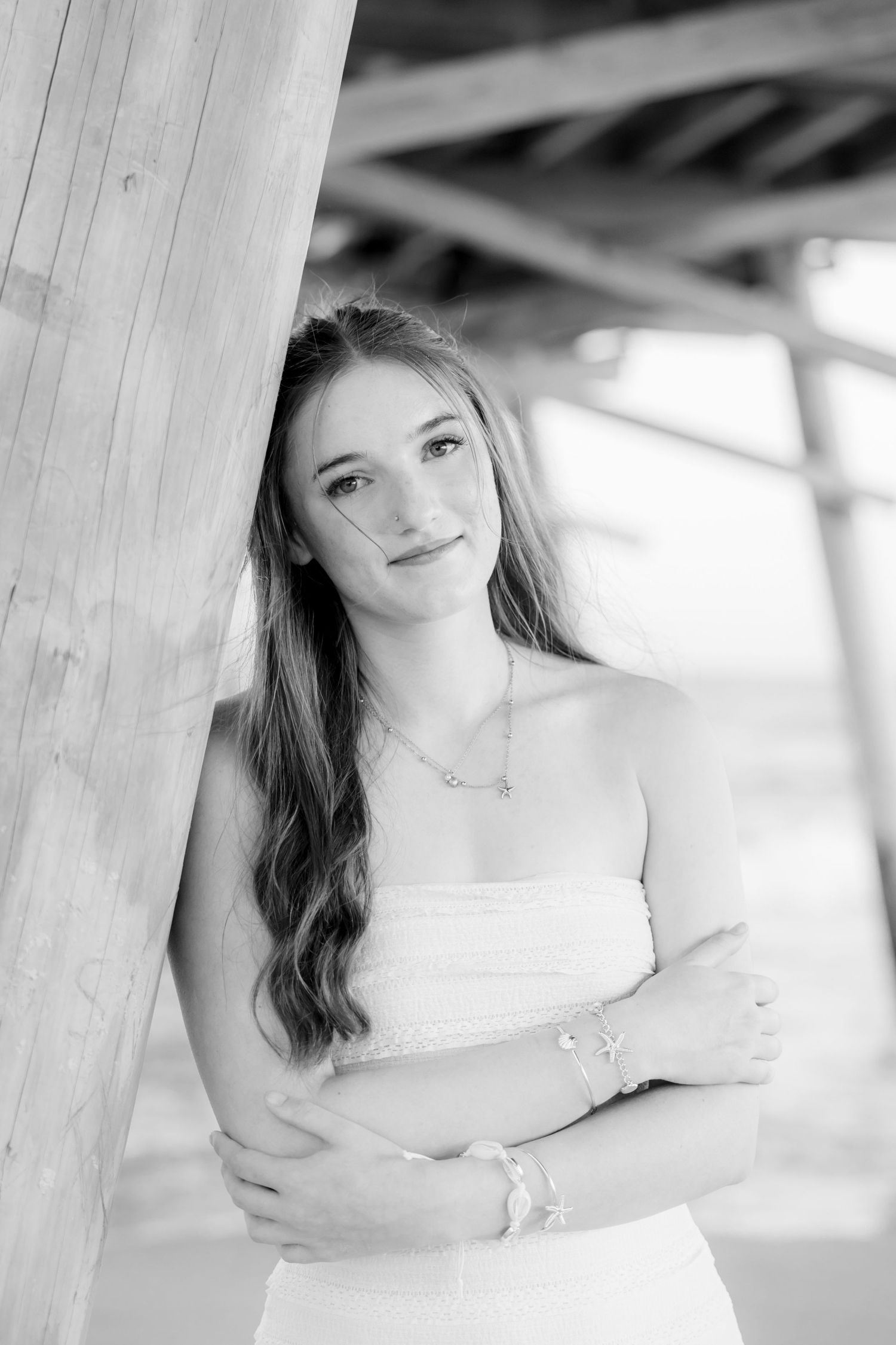 Black and white portrait of a young woman in a white dress leaning against a wooden post, arms crossed.