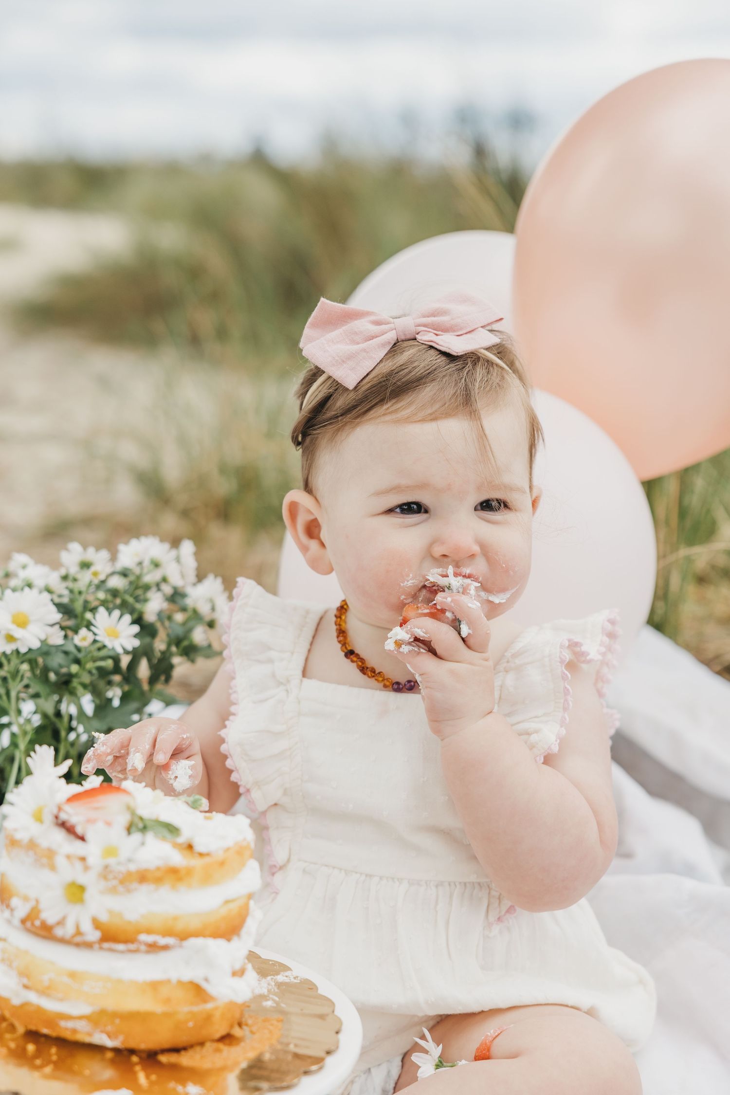 A baby in a white dress enjoys a cake smash photo session with pink balloons and flowers.