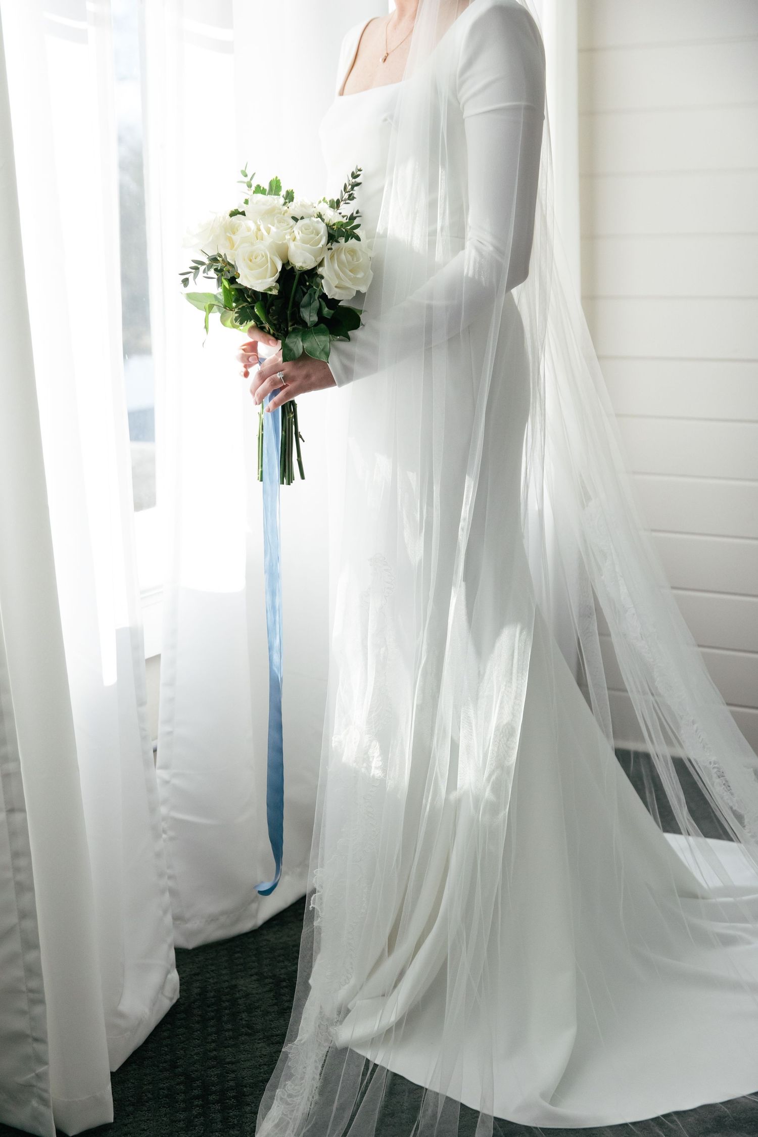 Elegant bride in long-sleeved white dress with flowing veil in natural light.