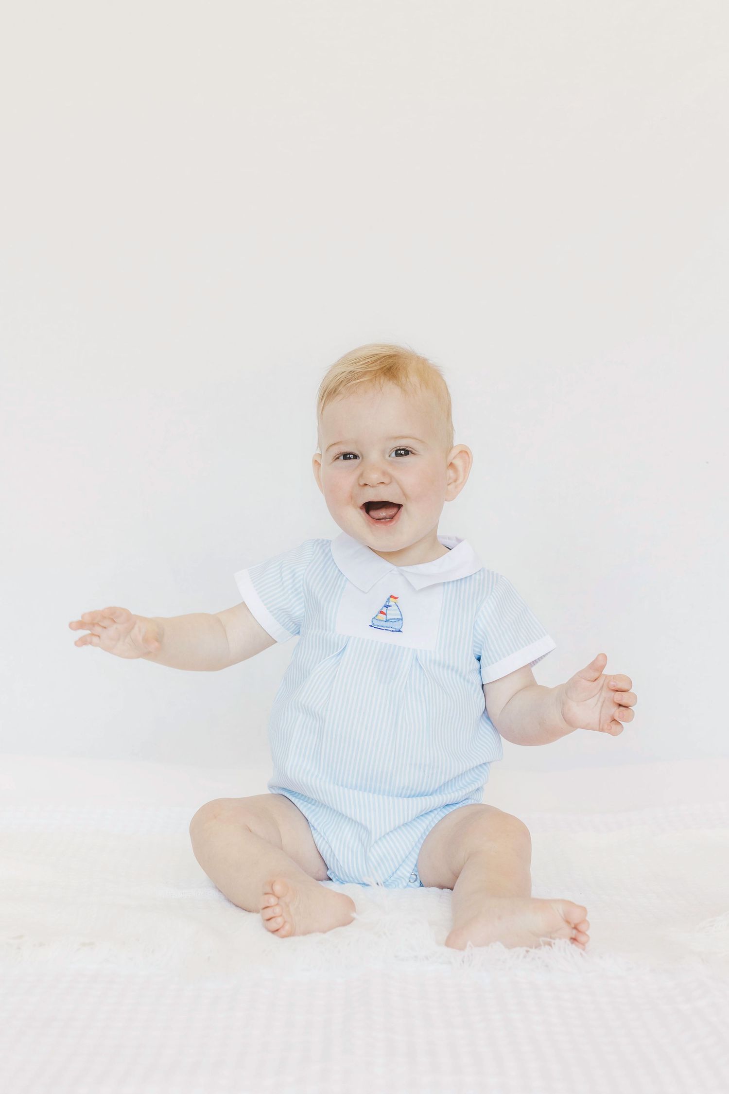 Happy baby wearing light blue romper sitting on white background with arms outstretched.