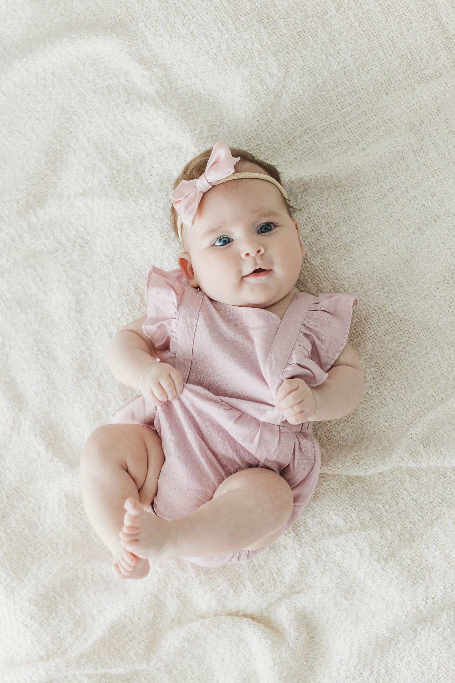 Baby in pink ruffle dress lying on soft white blanket.