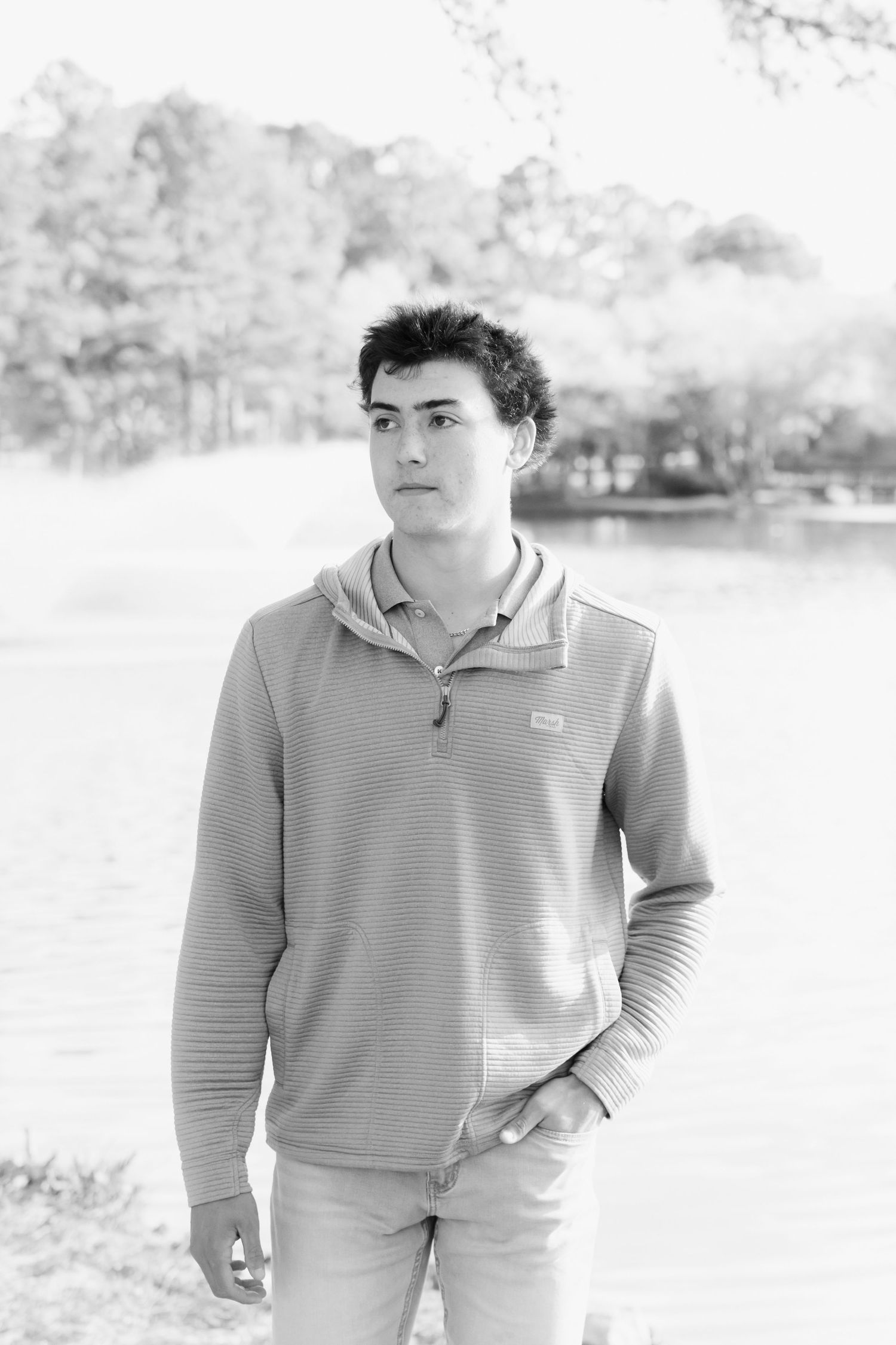 Black and white portrait of a teenage boy in a hoodie standing near a lake with trees in the background.