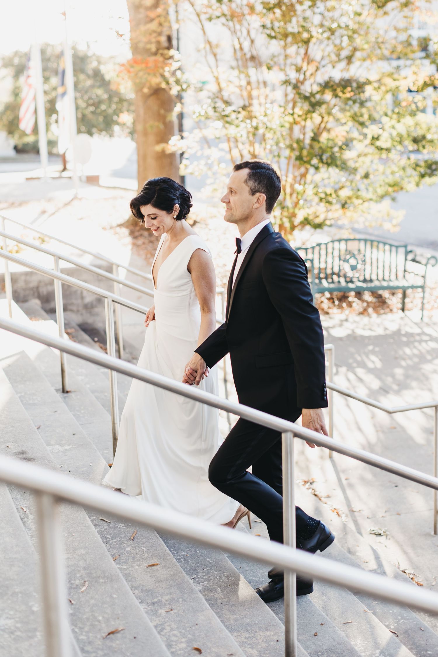 Couple in wedding attire walks up outdoor stairs holding hands.