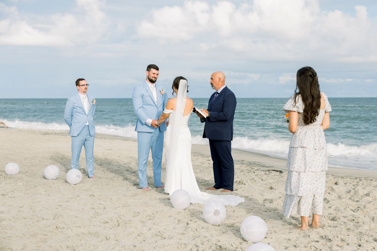 Beach wedding ceremony with light blue and white attire against ocean backdrop.