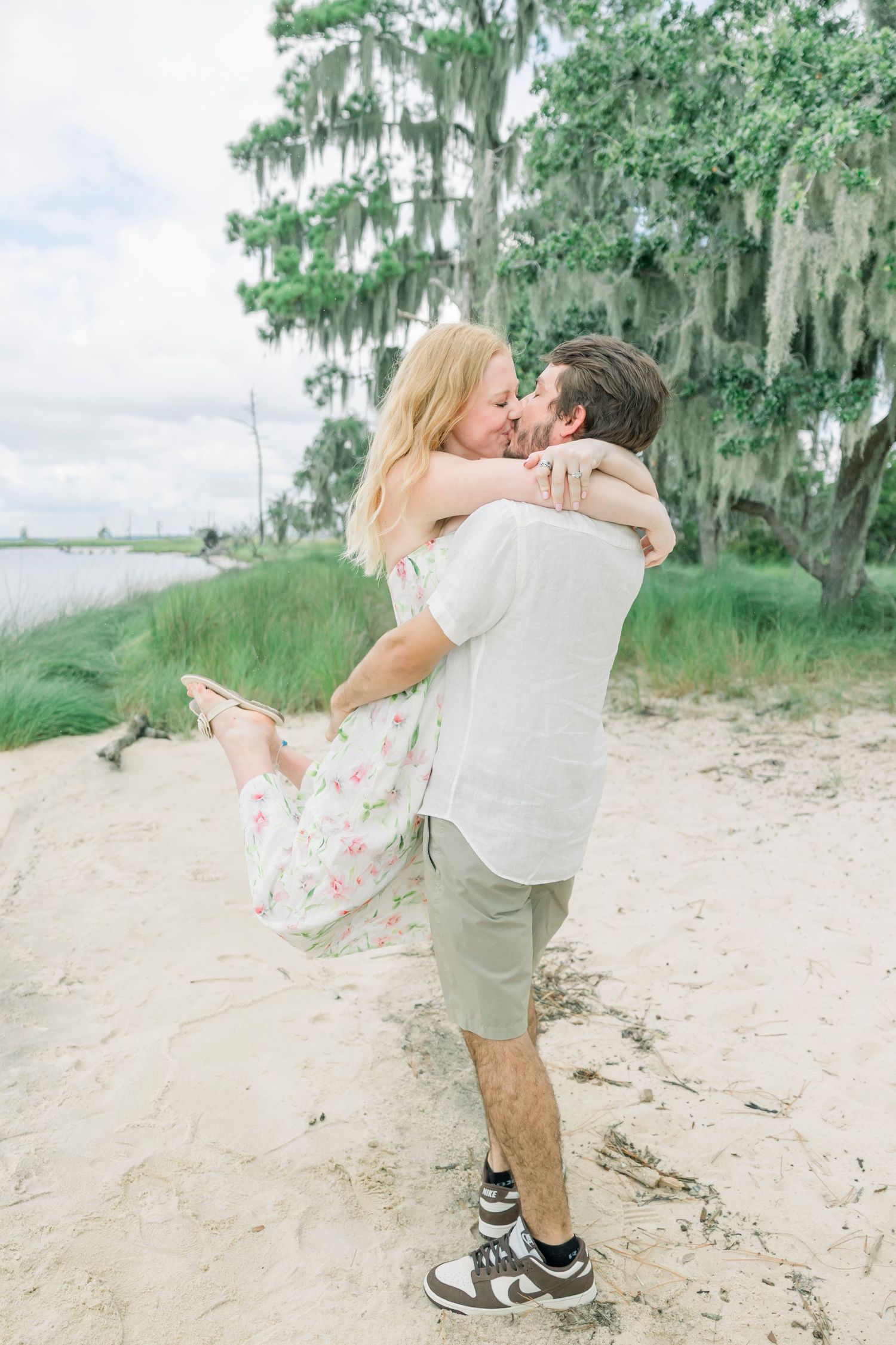 Couple embracing on a sandy beach with Spanish moss hanging from trees in the background.