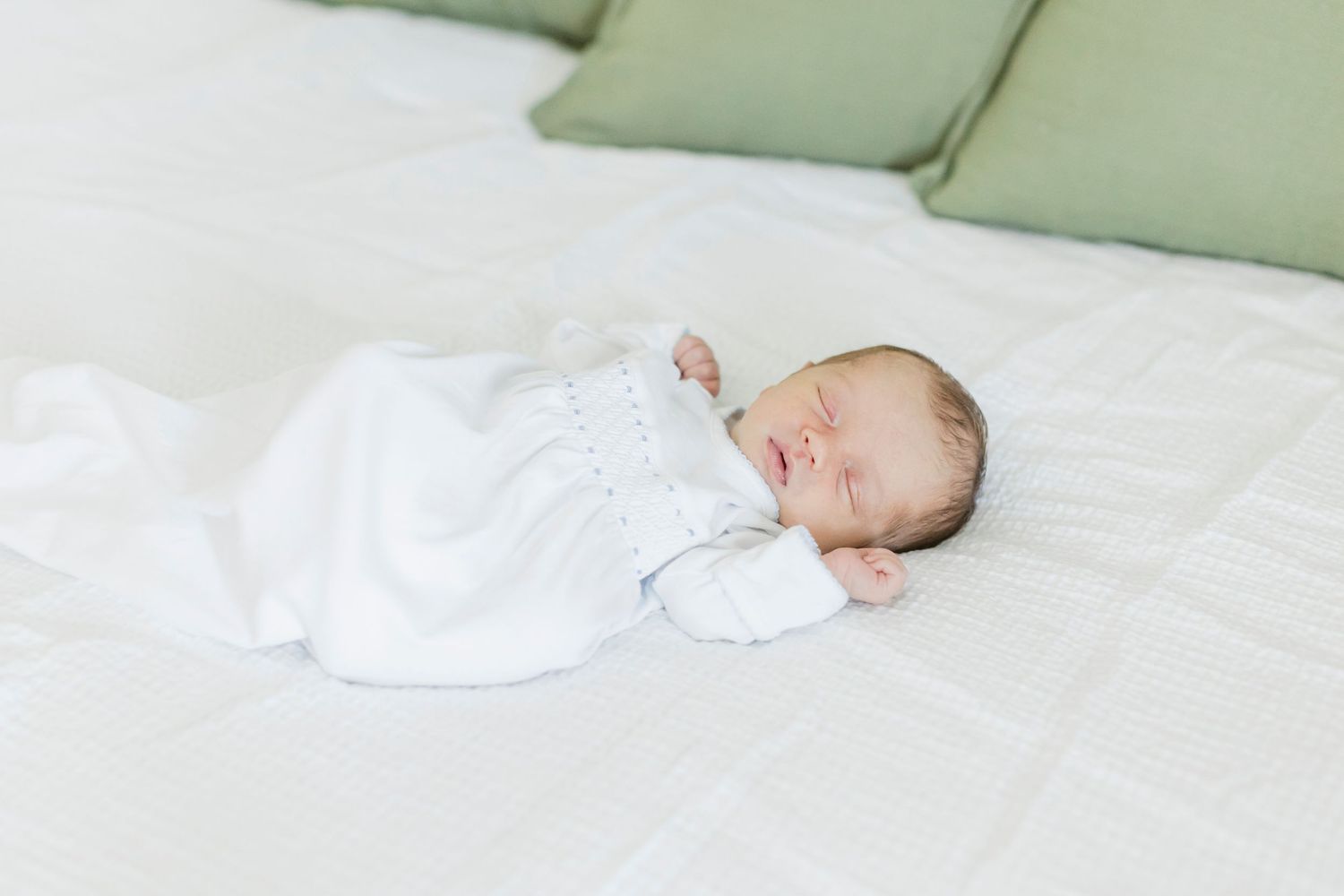 Peaceful sleeping infant wearing white sleepwear rests on crisp white bedding with soft green pillows.