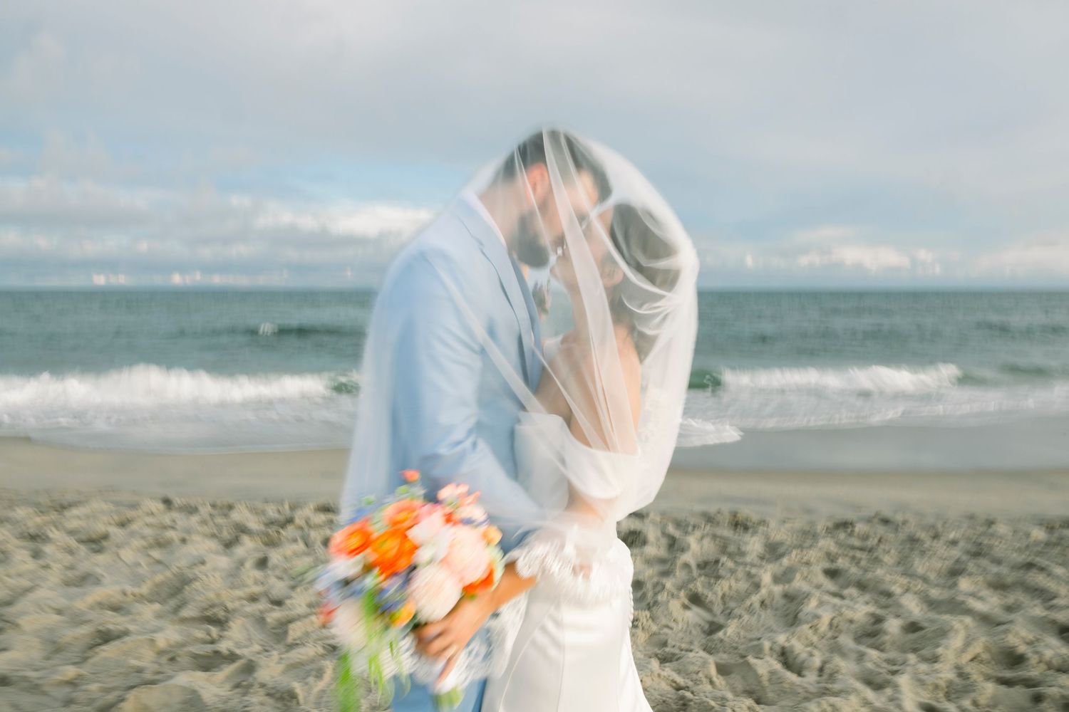 A couple embraces on a sandy beach with orange and pink flowers in the foreground.