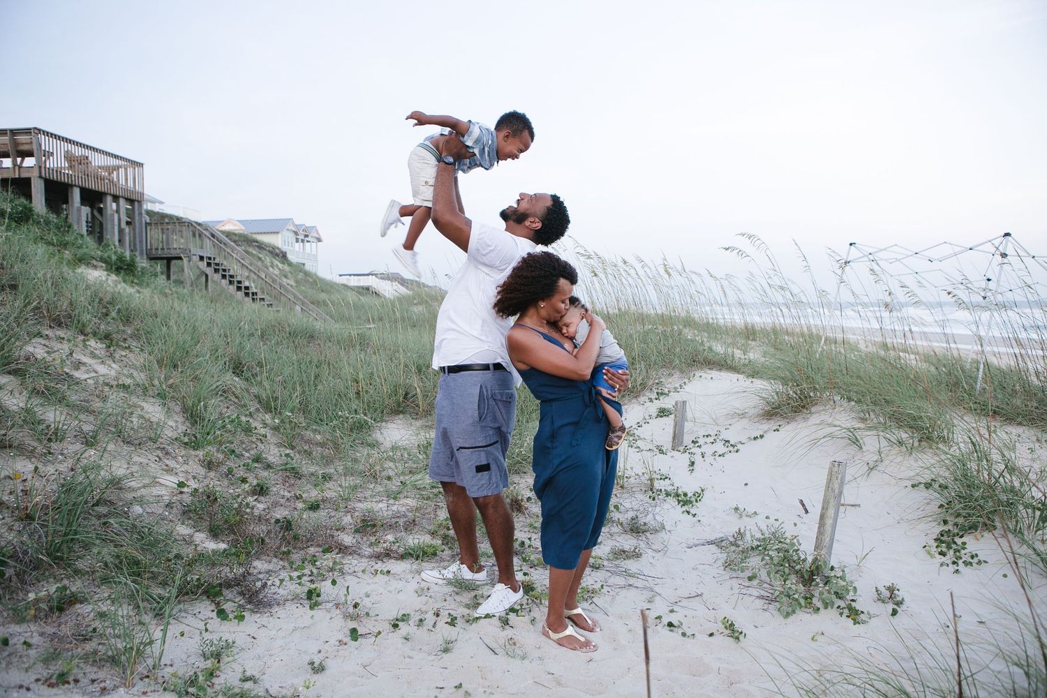 Family playing and lifting child in air on sandy beach dunes at sunset.