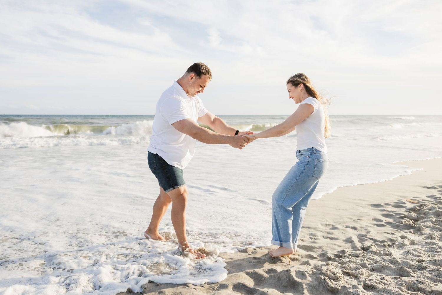 A playful couple holds hands while splashing in ocean waves on a sunny beach.