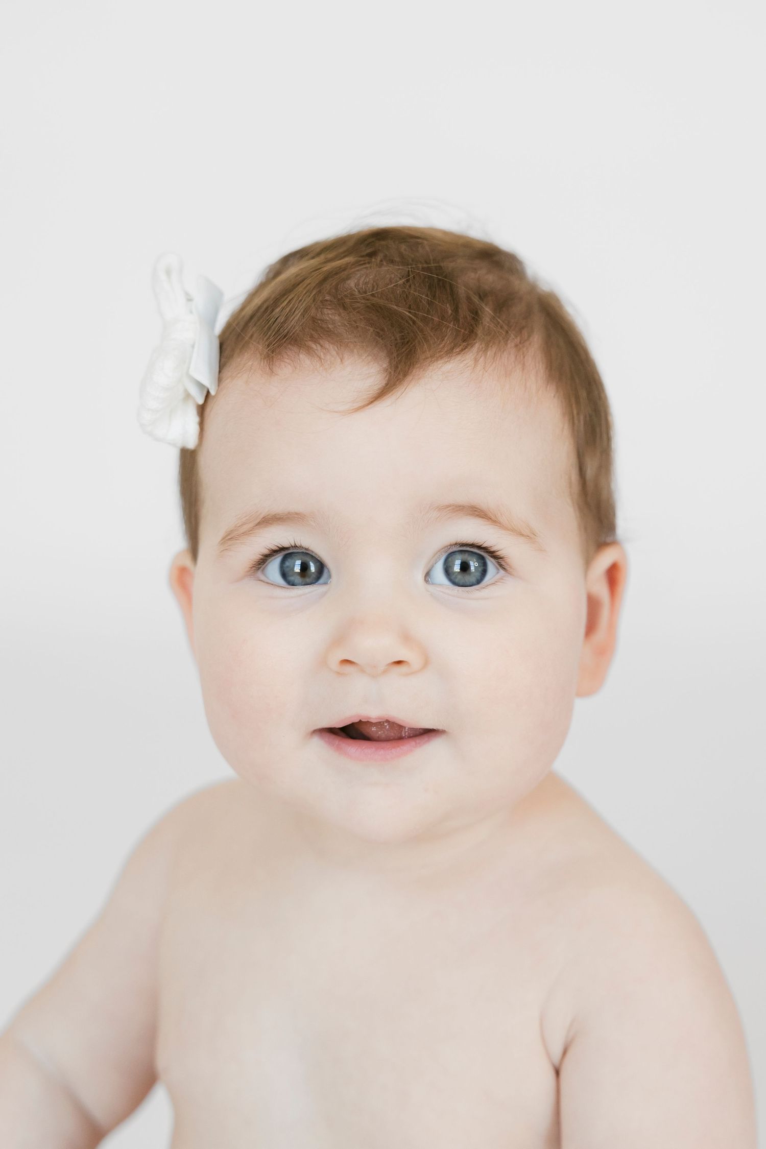 Baby wearing white bow accessory smiling against white background.