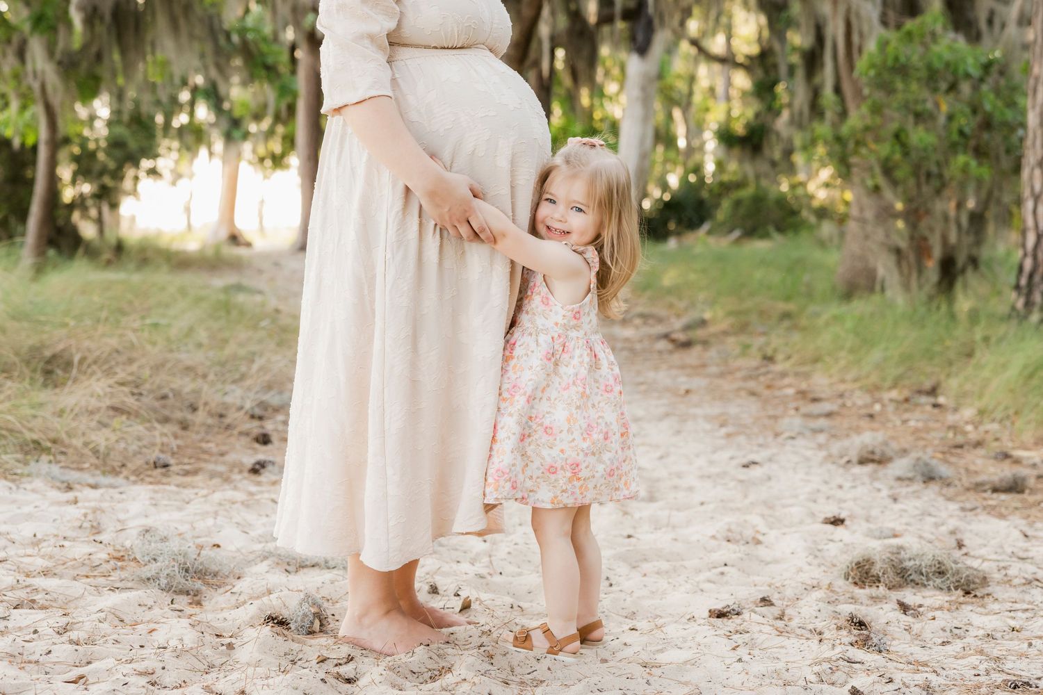 A young girl in a floral dress hugs her pregnant mother's belly outdoors on a sandy path surrounded by trees.