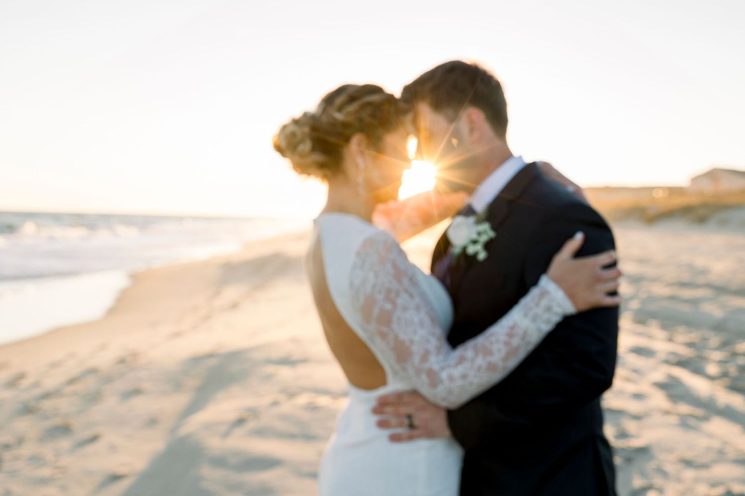 Romantic beach wedding photoshoot at sunset with couple embracing on sandy shore.
