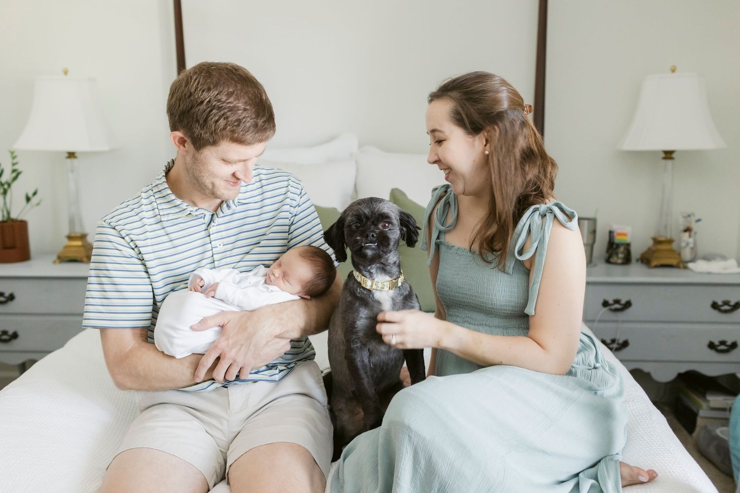 Family sits on bed with their black dog while holding a newborn in a cozy bedroom setting.