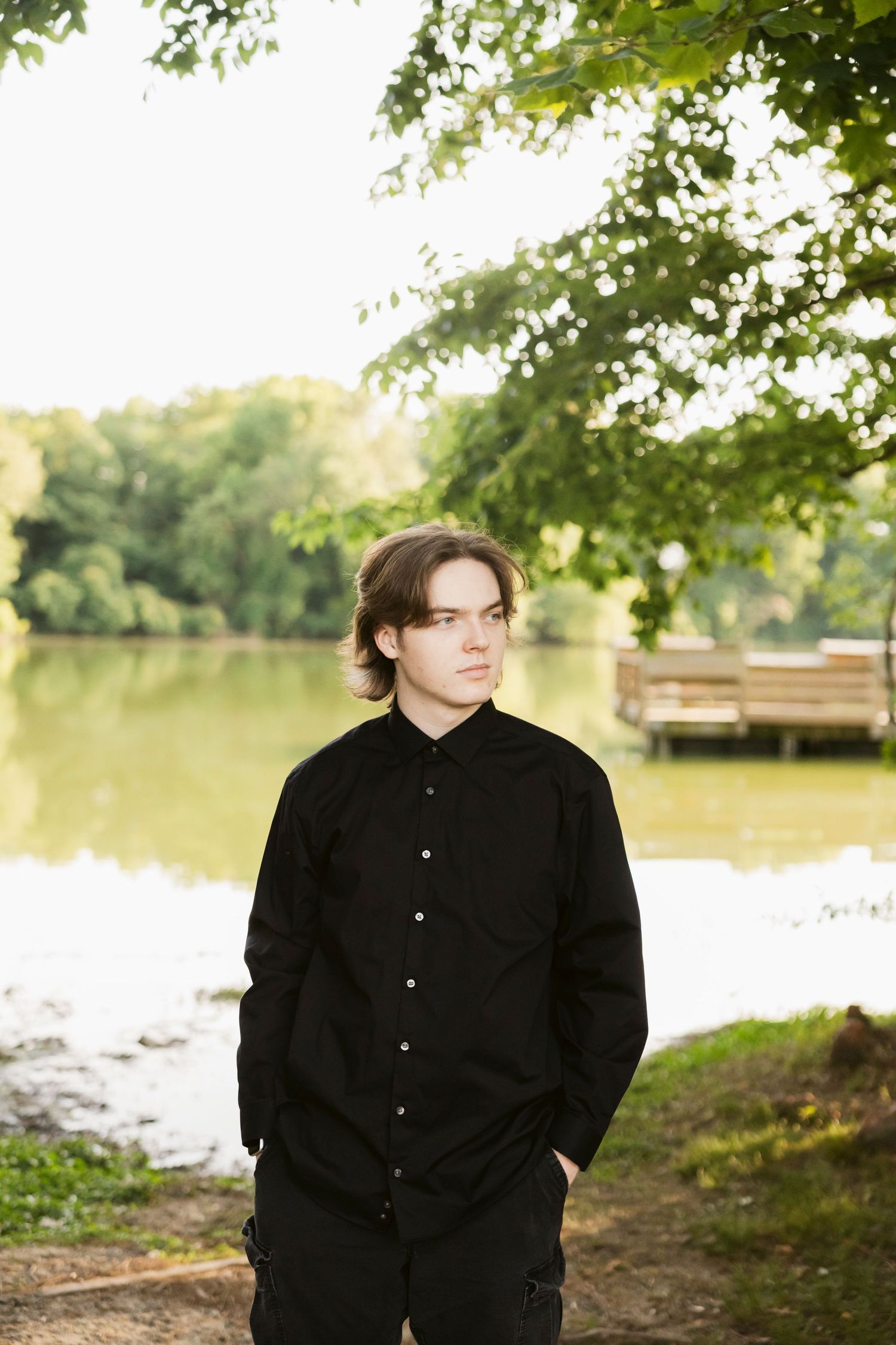 Young man in black shirt and pants standing outdoors near a calm lake surrounded by lush green trees.