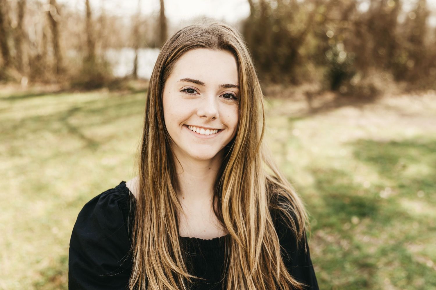 Portrait of someone with long brown hair smiling outdoors with natural lighting.
