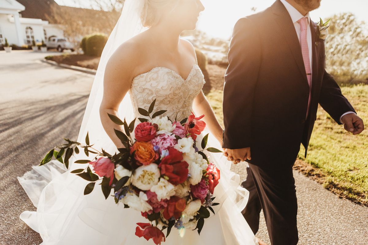 Sunlit wedding couple holding vibrant red and pink bouquet outdoors.