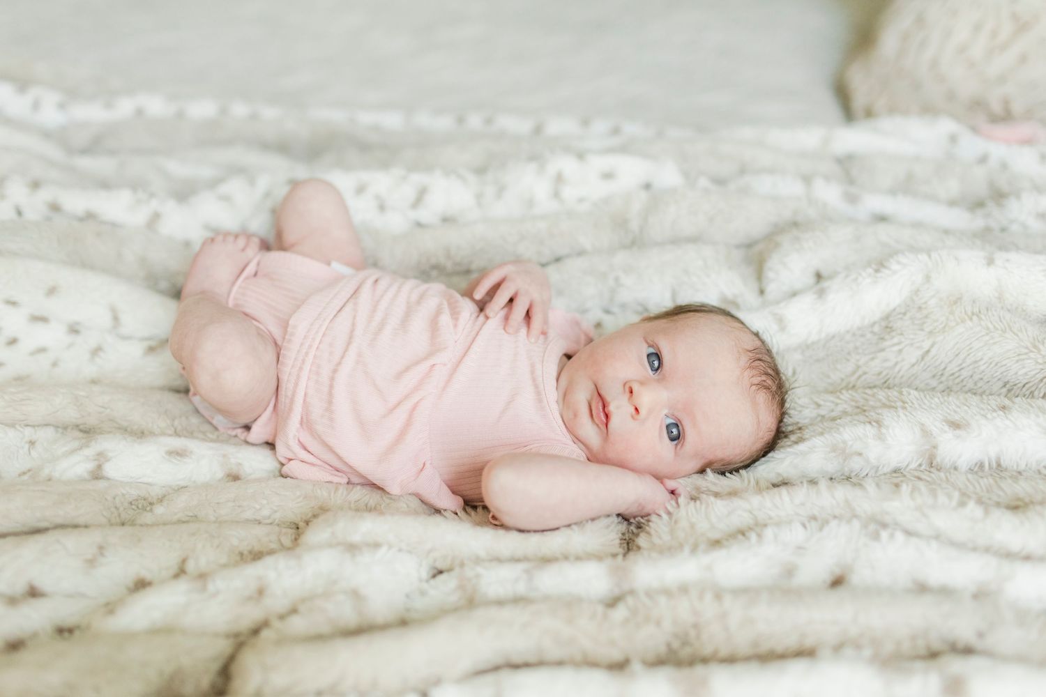 Baby in pink onesie lying on textured white blanket looking at camera.
