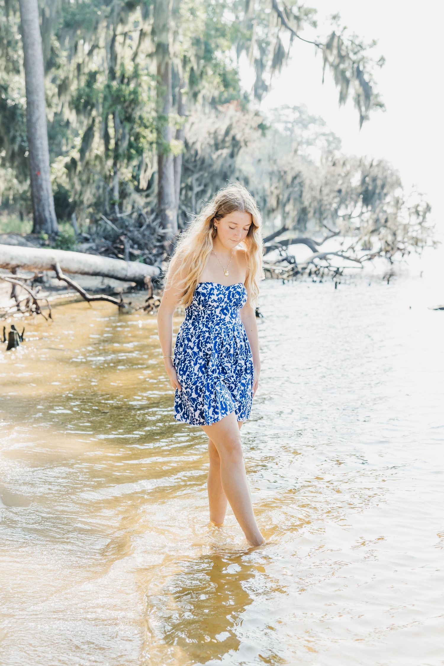 Woman in blue floral dress splashes in shallow coastal waters near moss-covered trees.