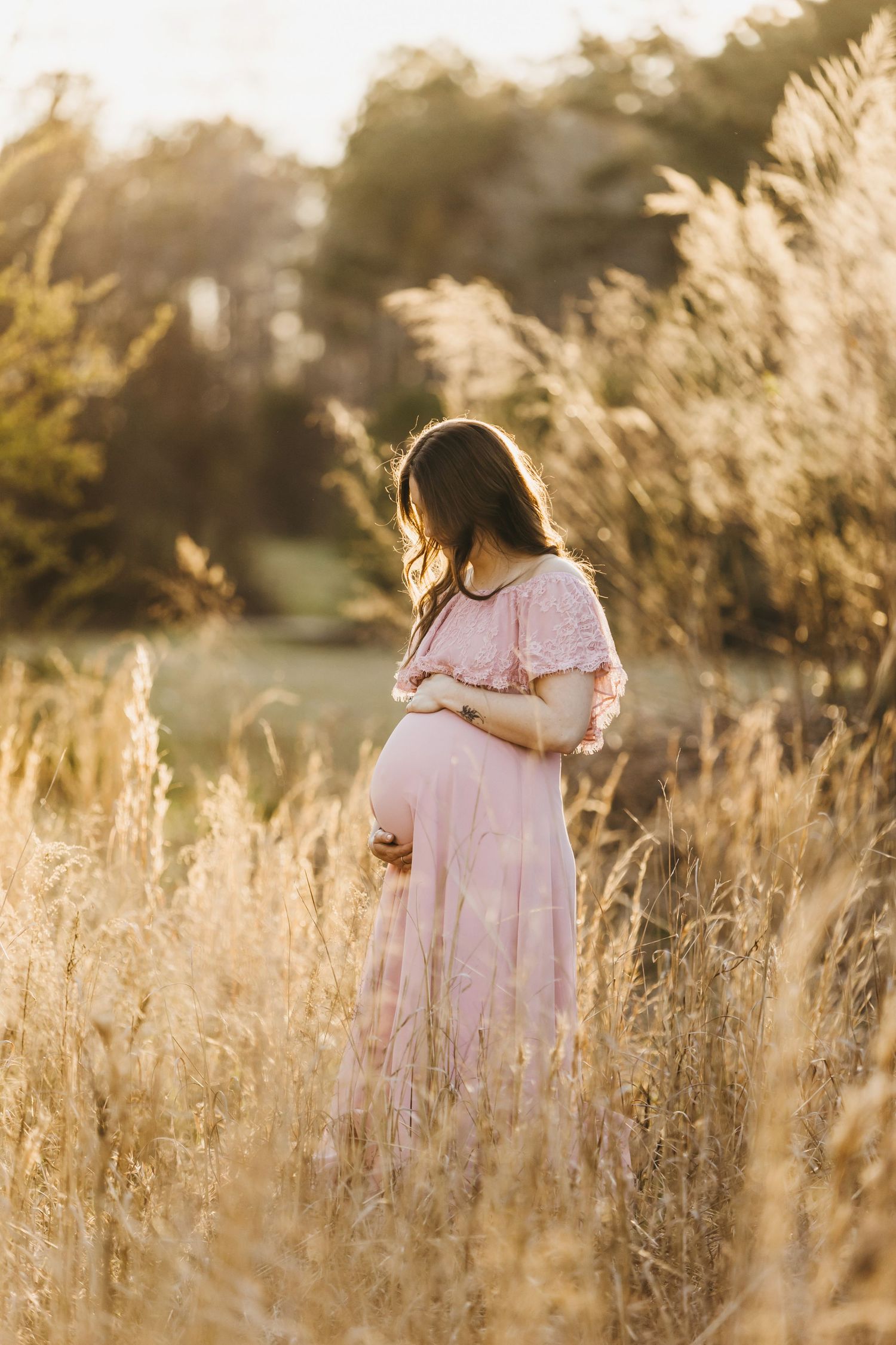 Person in pink dress poses in golden field during sunset maternity photography session.