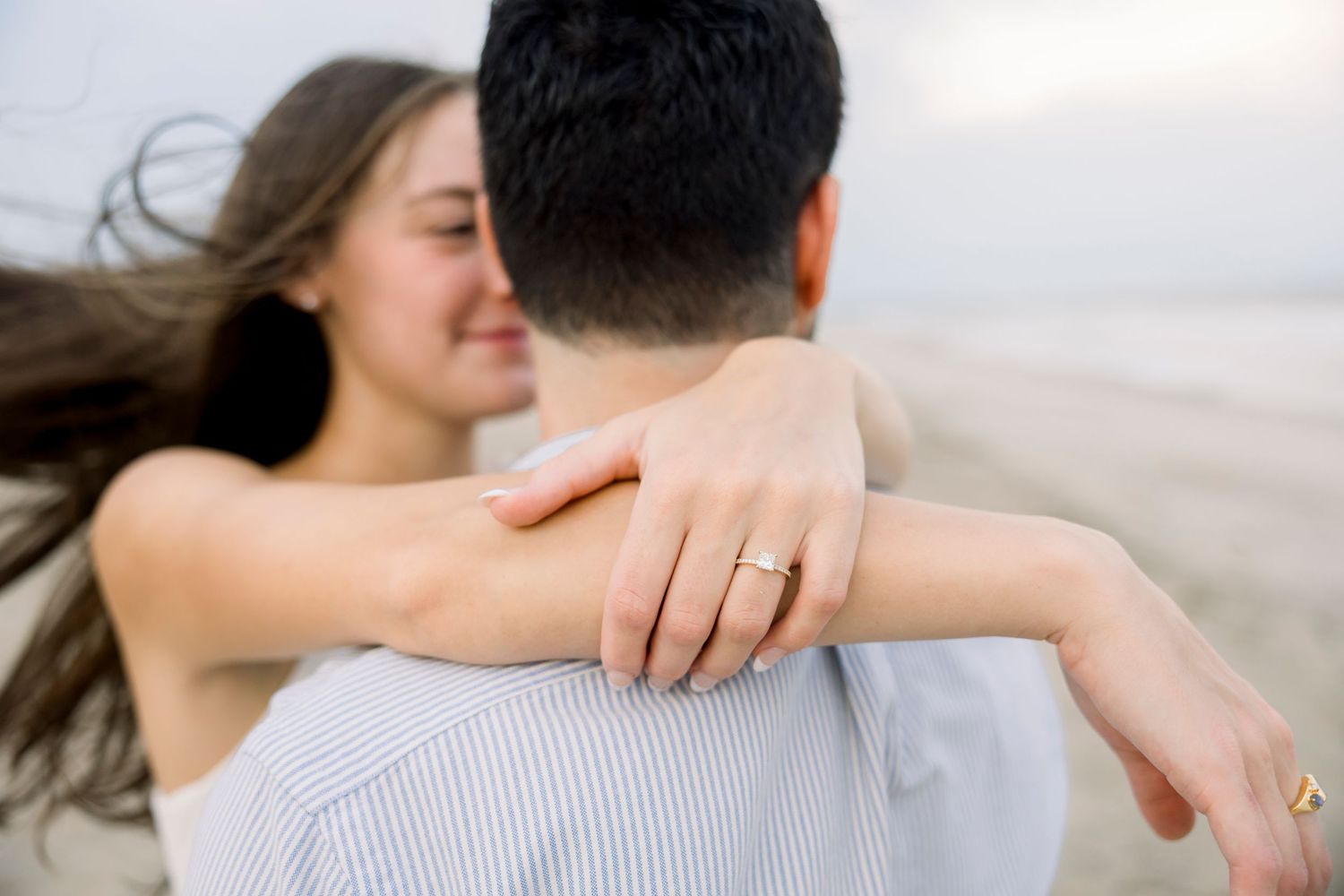 Couple embracing on beach, woman wearing gold engagement ring, hair blowing in wind.