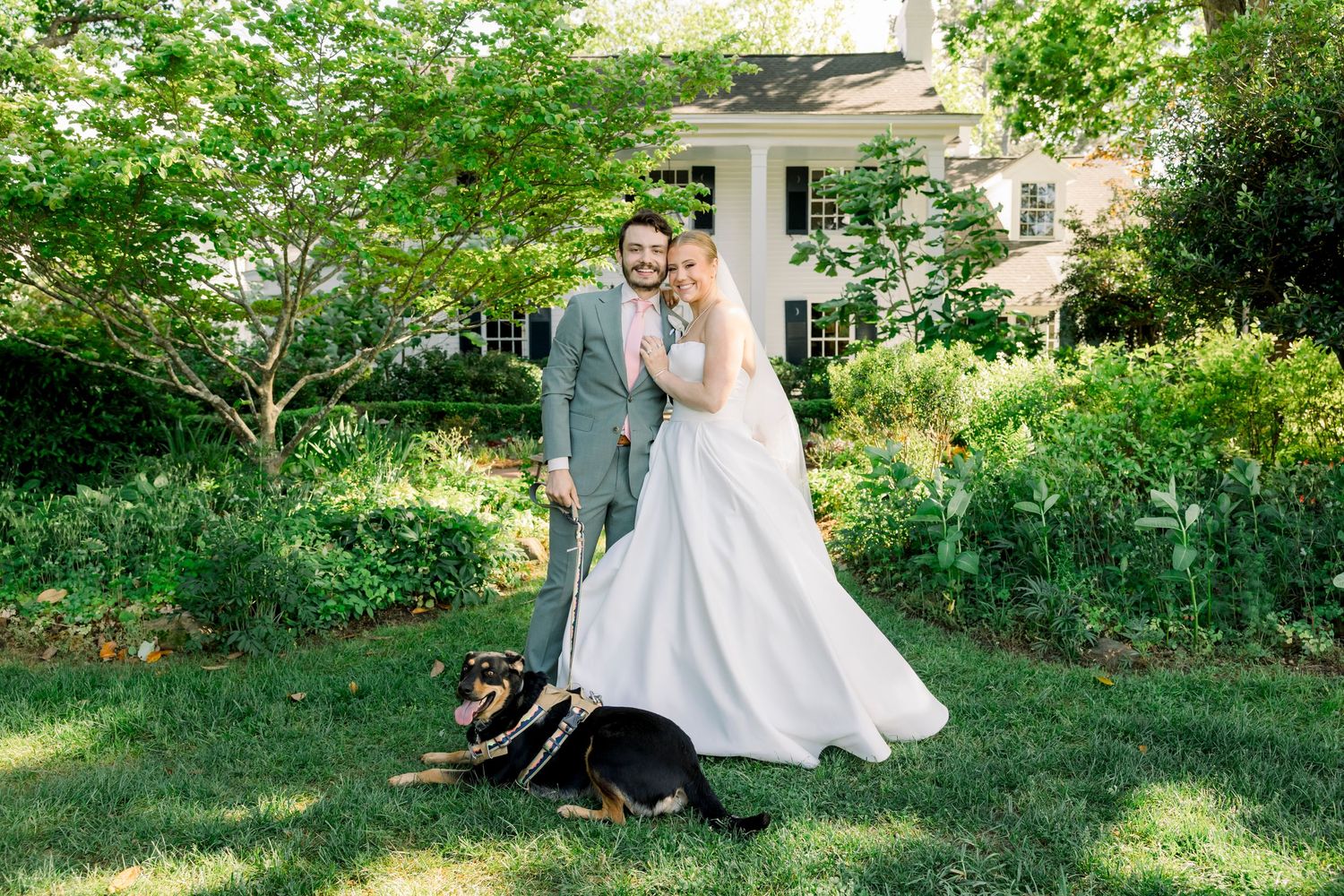 Bride and groom pose with their dog in a lush garden outside a white house on their wedding day.