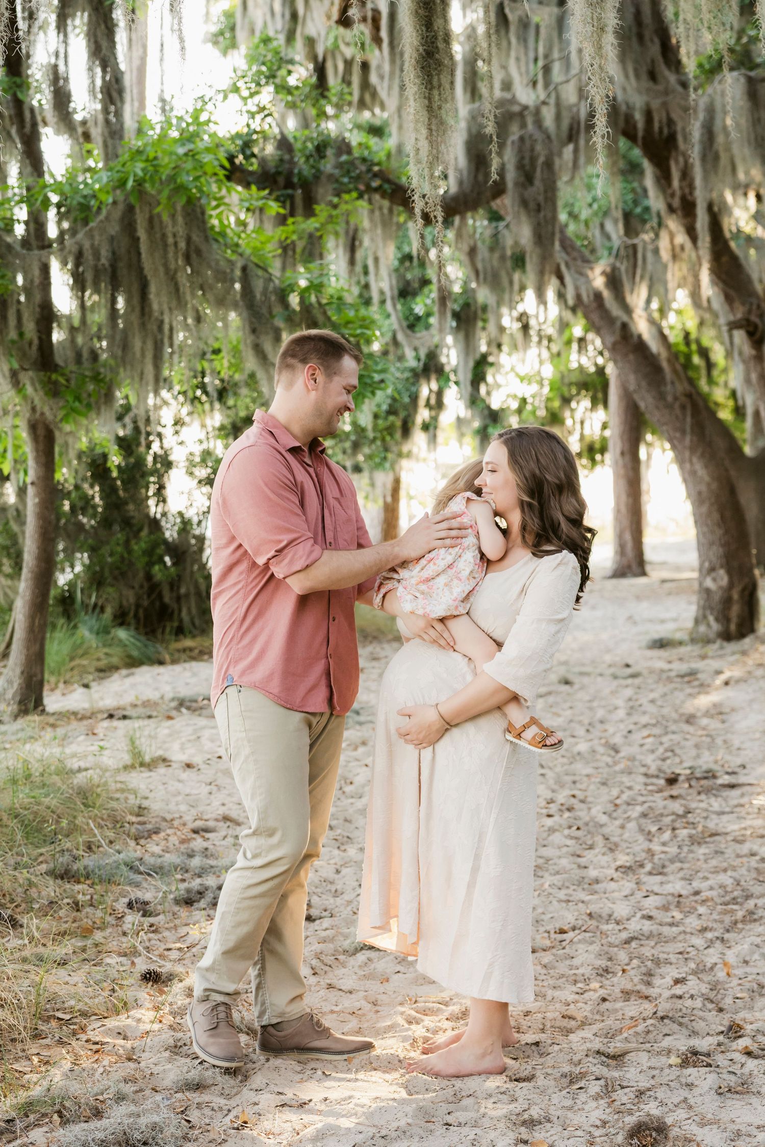 A couple shares a tender moment outdoors under moss-draped trees, the woman in a white dress cradling her baby bump.