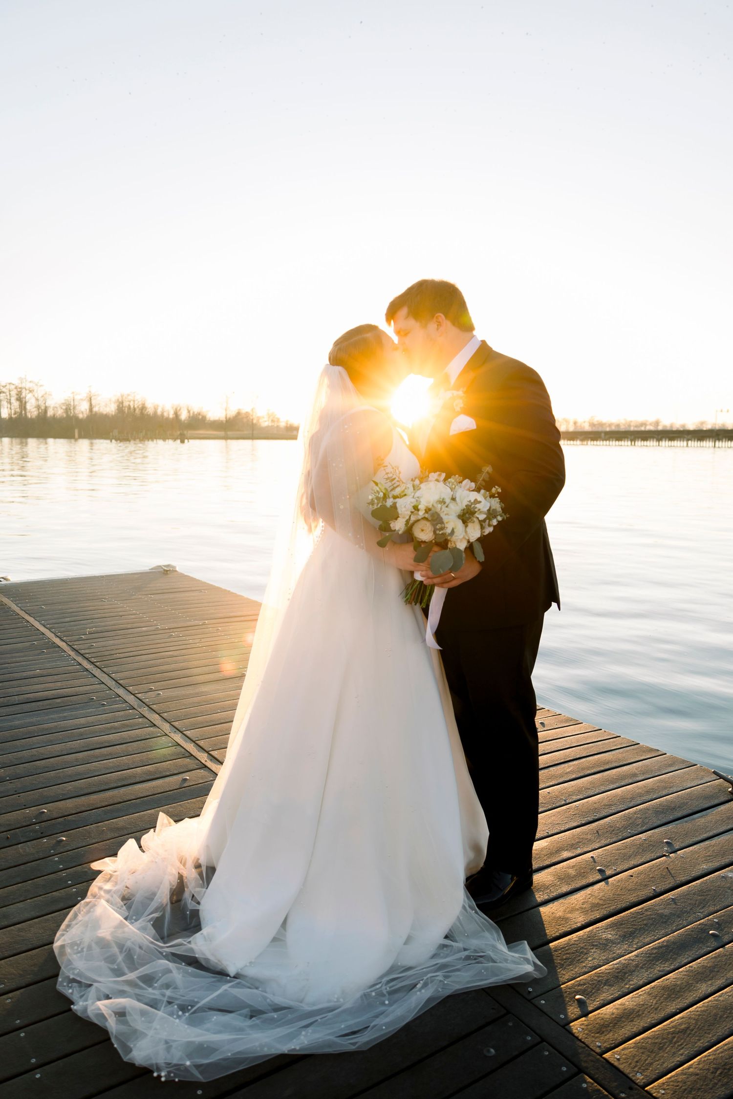 Romantic sunset silhouettes of a couple in wedding attire embracing on a wooden dock by the water.