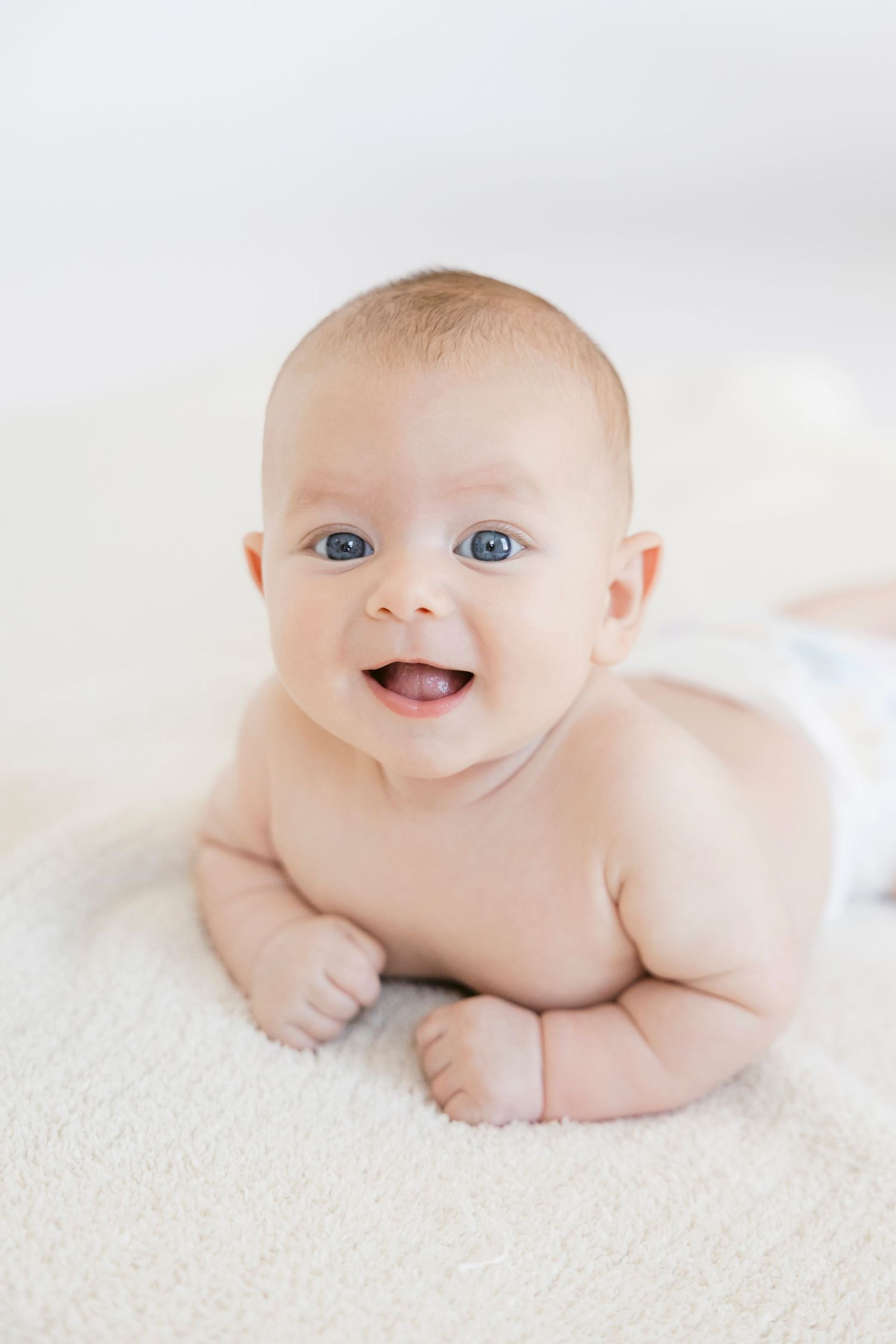Adorable smiling baby with blue eyes doing tummy time on a soft white blanket.