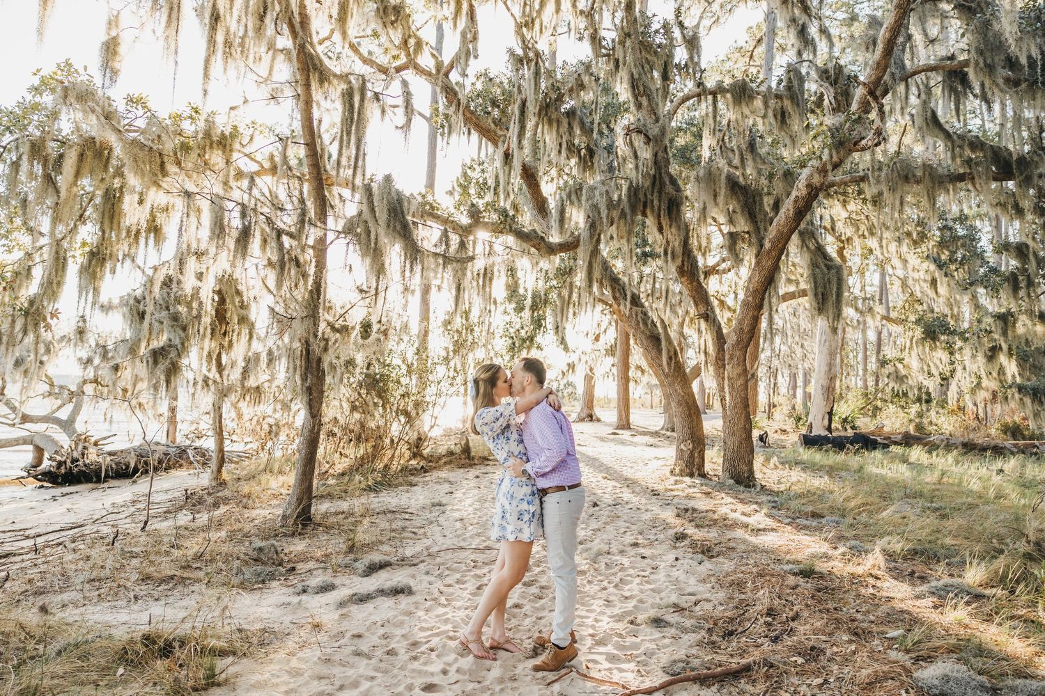 A couple kissing and embracing under Spanish moss hanging in trees along a serene coastal pathway.