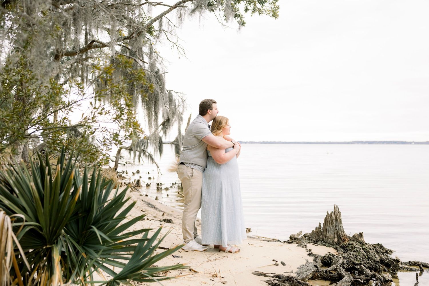 A couple embraces on a serene beach with palm trees and Spanish moss.
