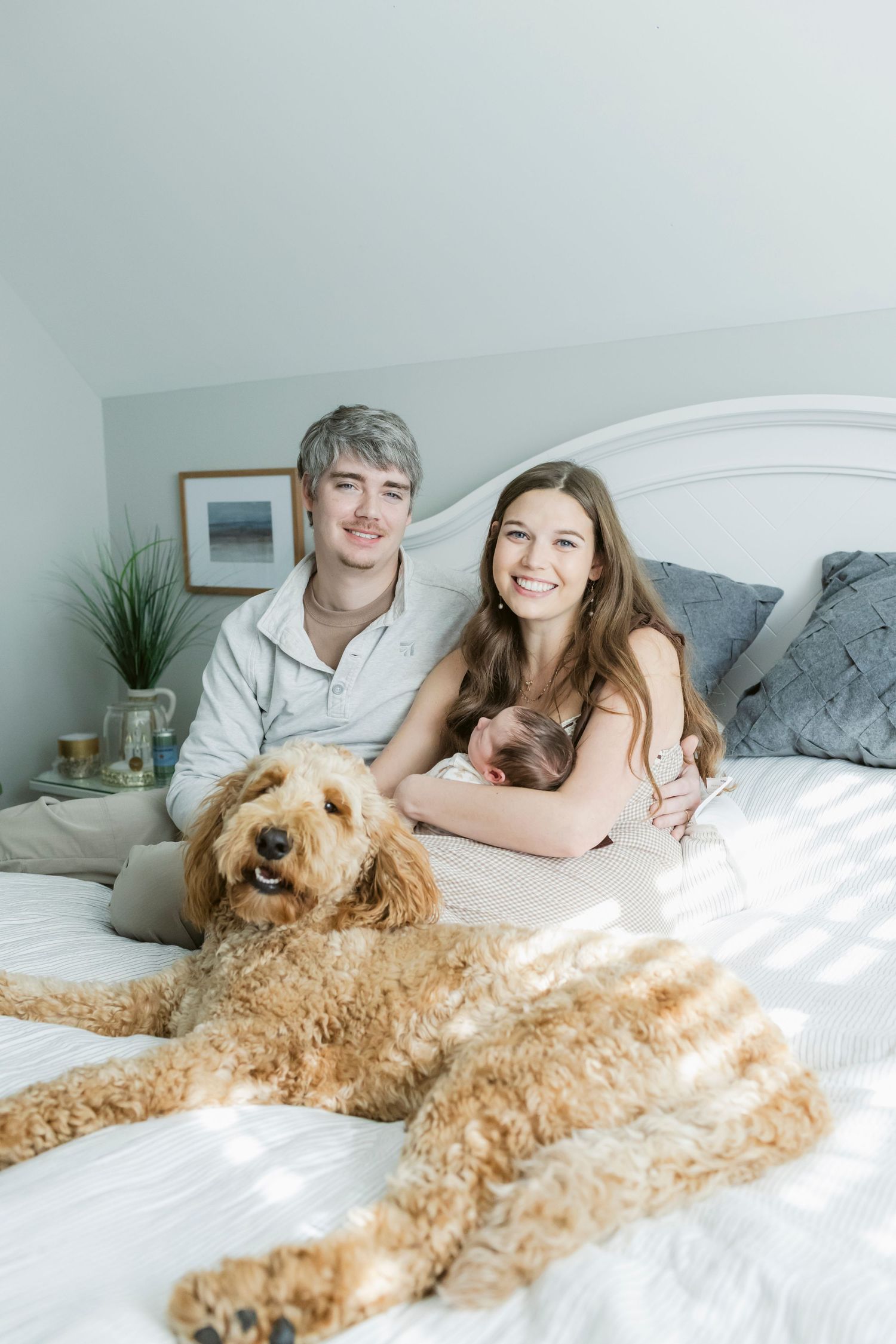 A goldendoodle dog lounges on a white bed with its owners in a bright bedroom setting.