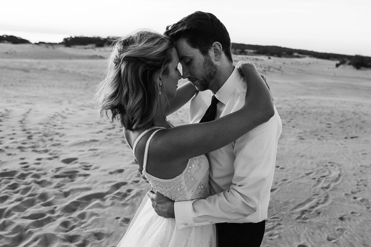 A romantic couple embraces on a sandy beach during their wedding photoshoot at sunset.