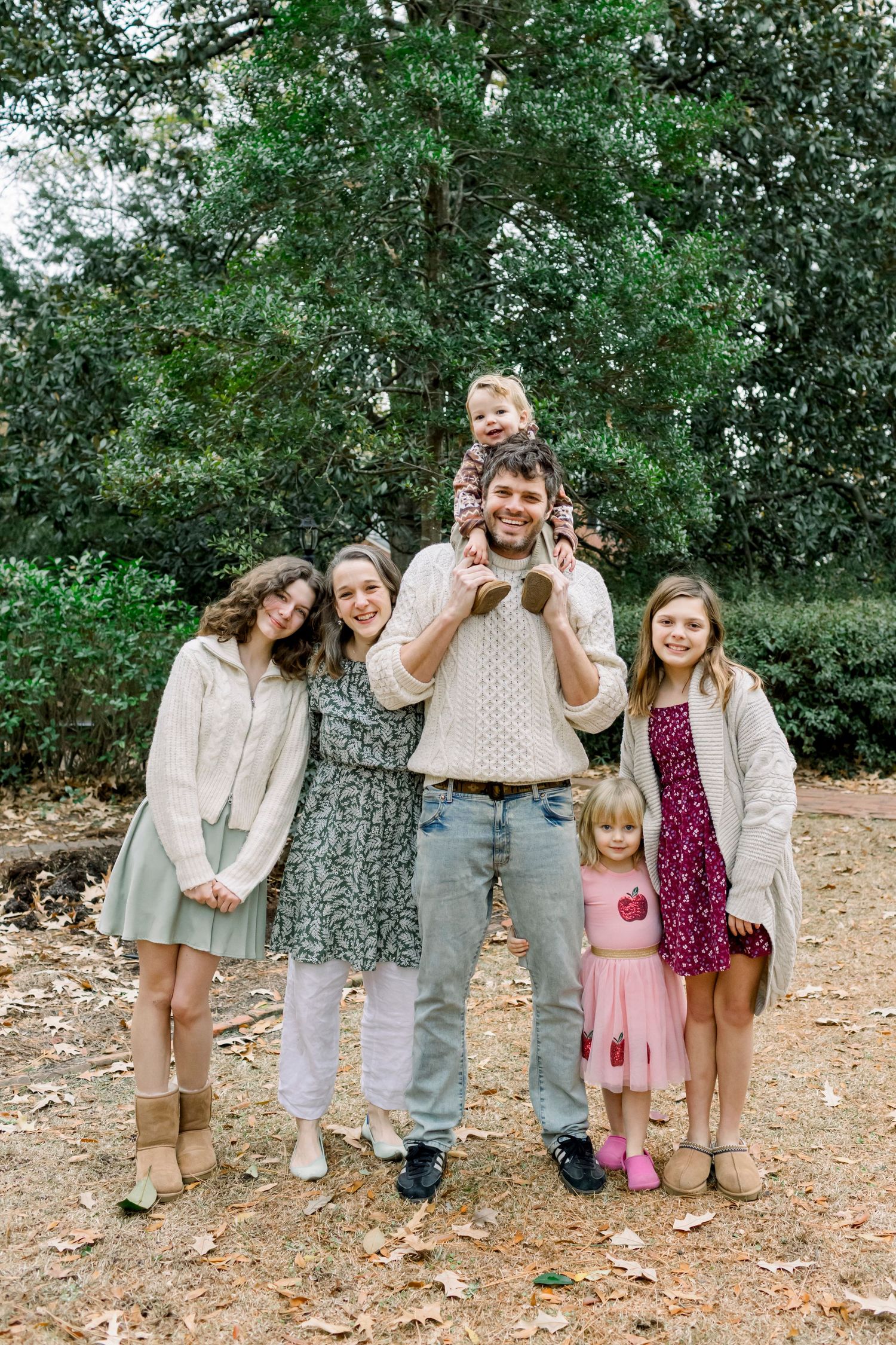 Family group wearing coordinated neutral tones poses together in outdoor woodland setting.
