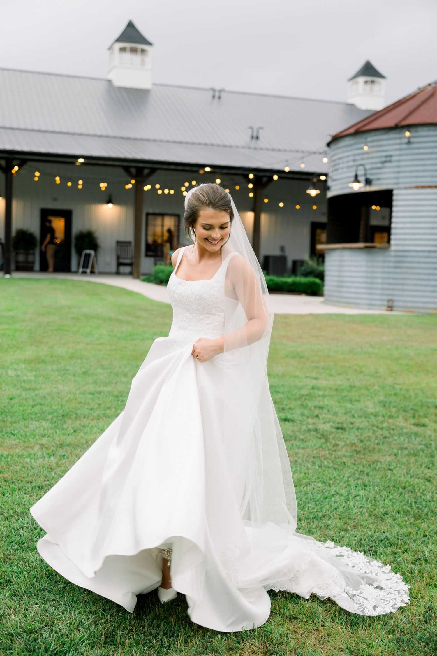 A flowing white wedding dress twirls on a grassy lawn near white farm buildings.