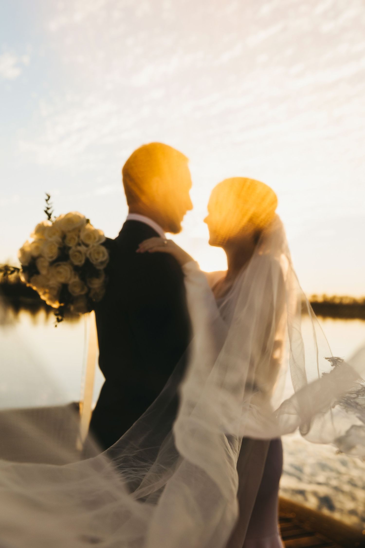Silhouetted wedding couple against sunset sky with flowing veil and bouquet.
