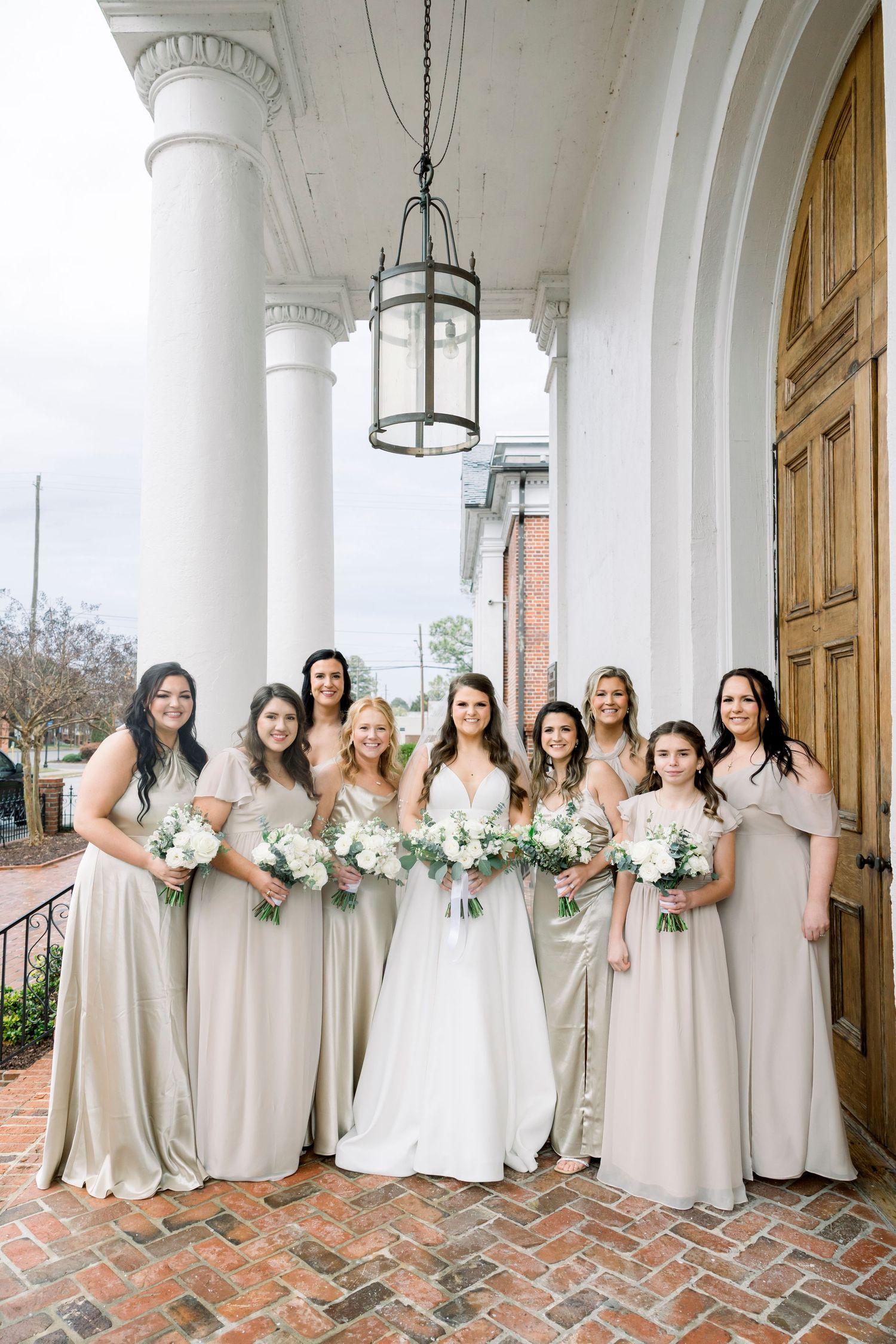 Bridesmaids in light neutral dresses pose with bouquets under white columned portico.
