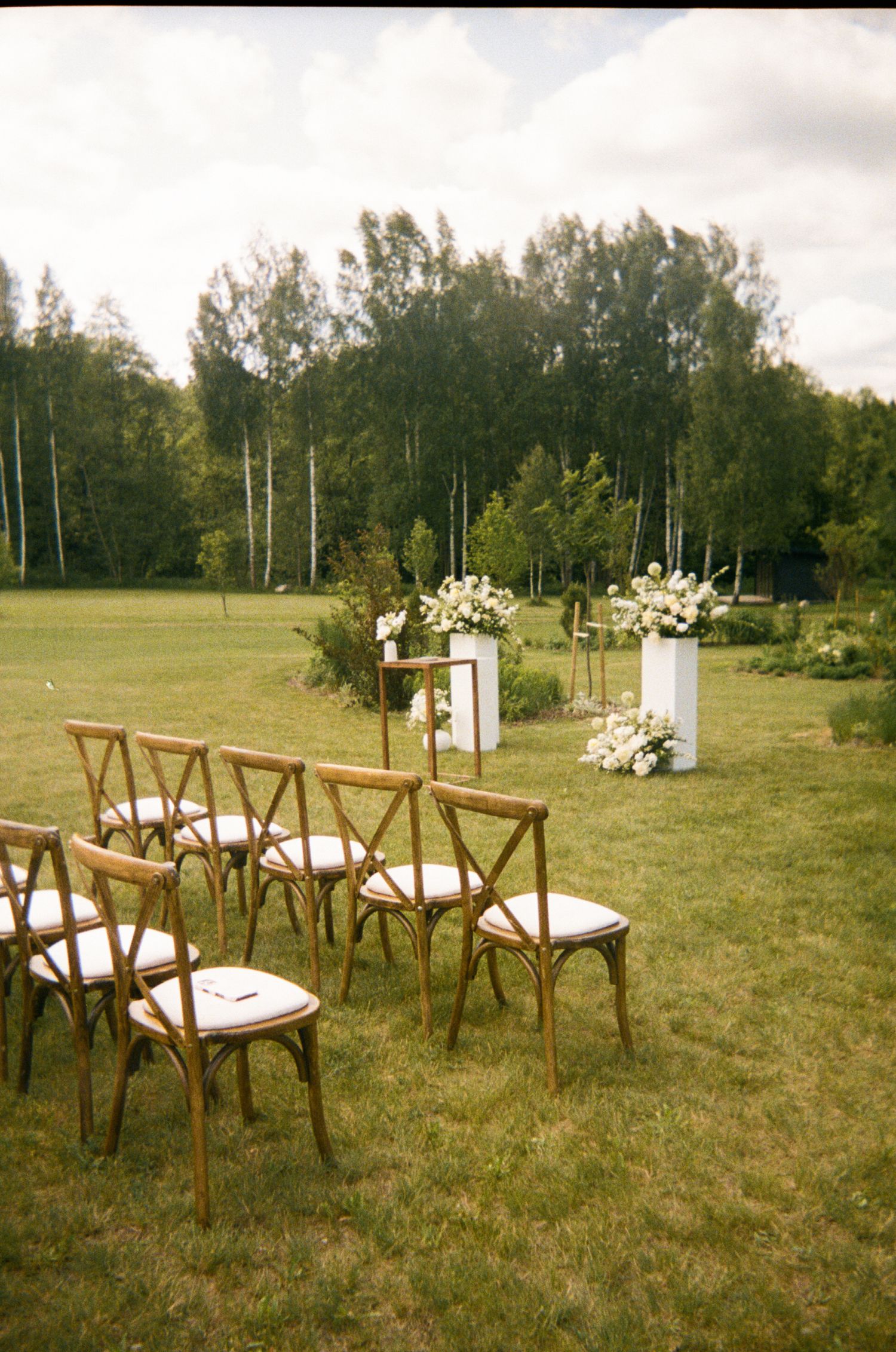Wooden crossback chairs set up on grass for an outdoor ceremony with white floral decorations.