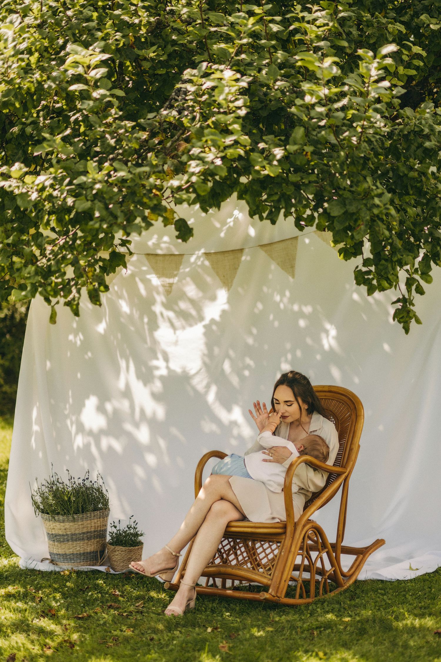 A woman sits in a rattan rocking chair outdoors, cradling a baby beneath a lush tree with a white backdrop.